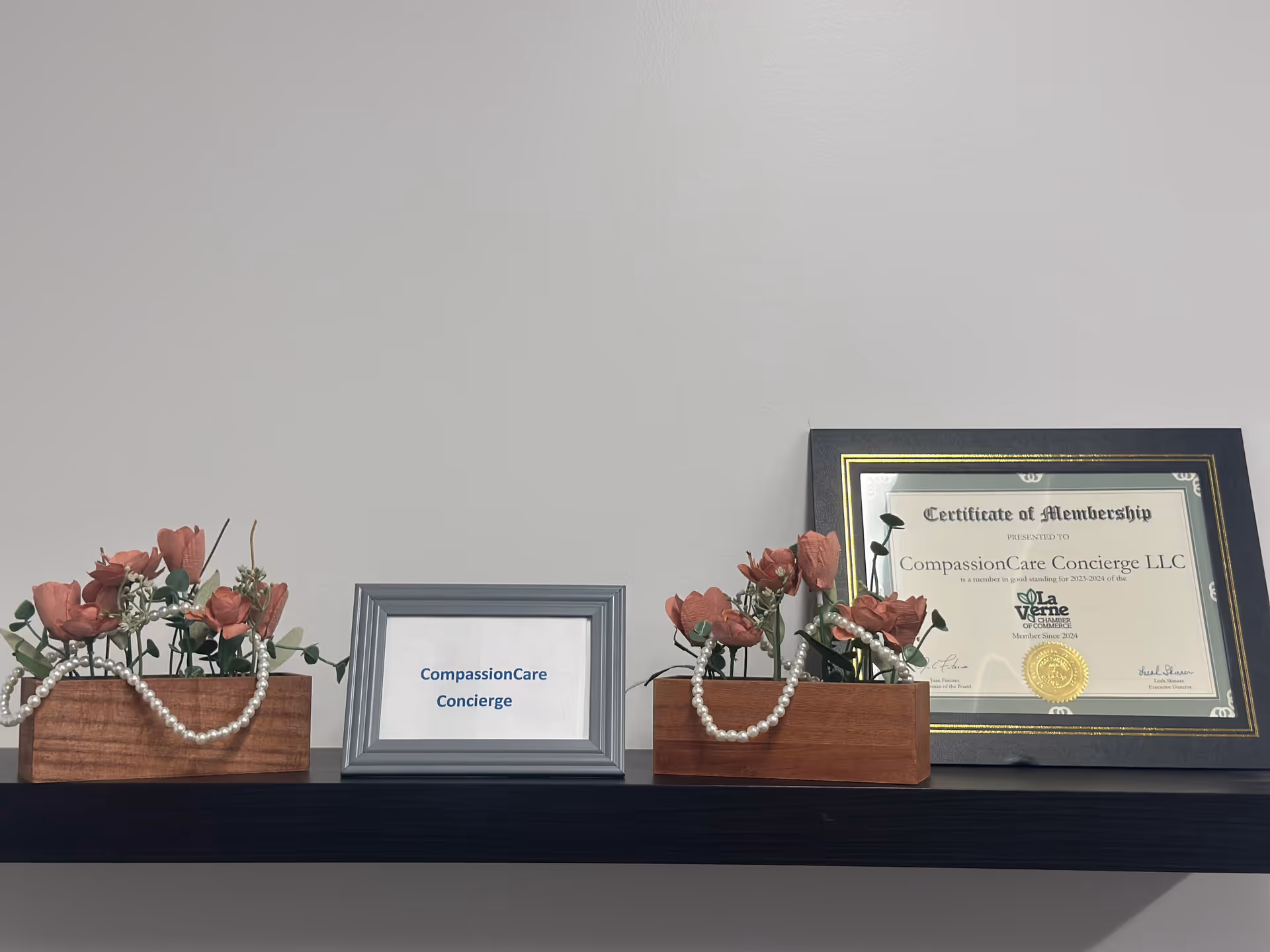 Shelf with flower arrangements, pearl strands, and a framed CompassionCare Concierge certificate.