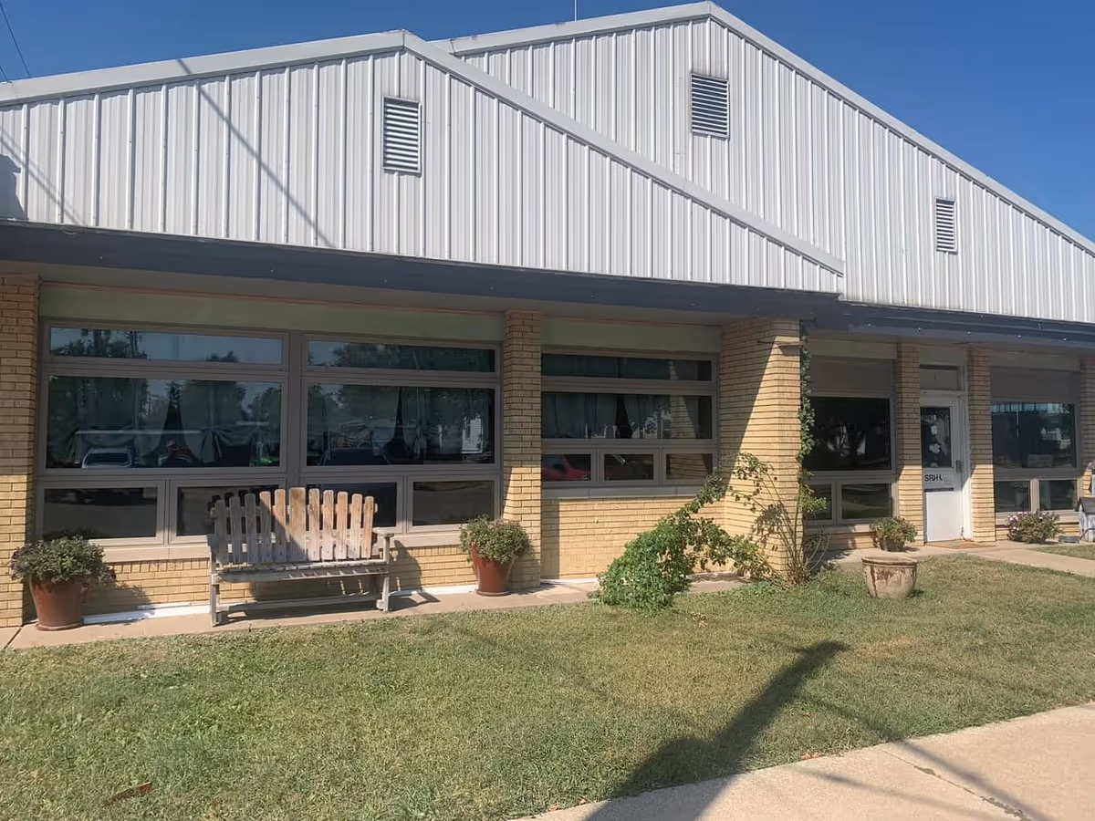 Brick facility exterior with large windows, a bench, and potted plants at the entrance.