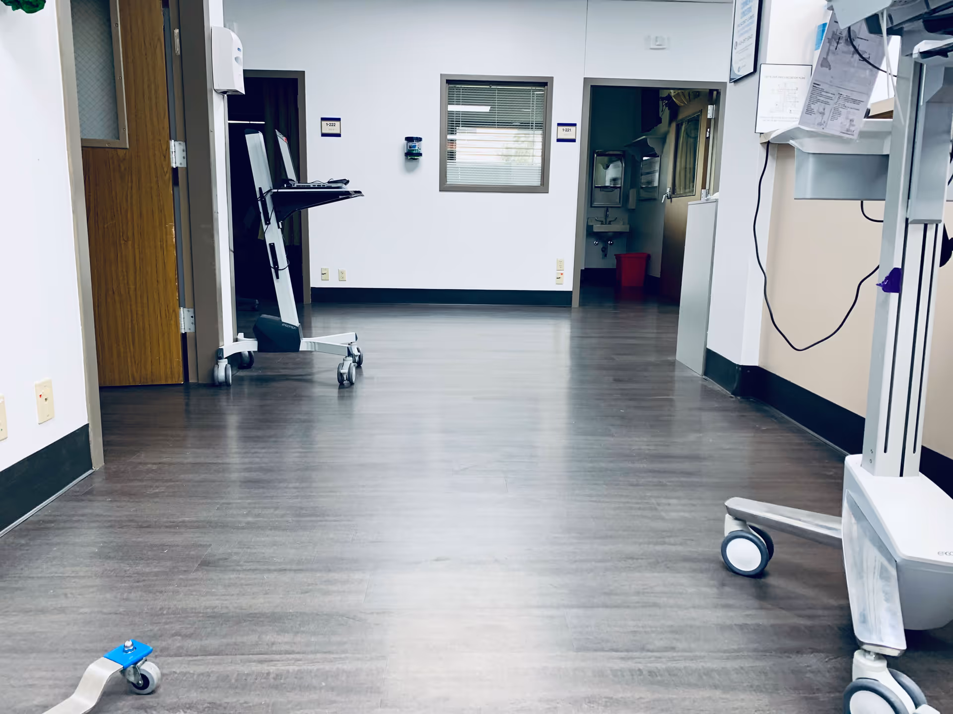 A sterile hospital hallway with medical equipment carts and a doorway at the far end.