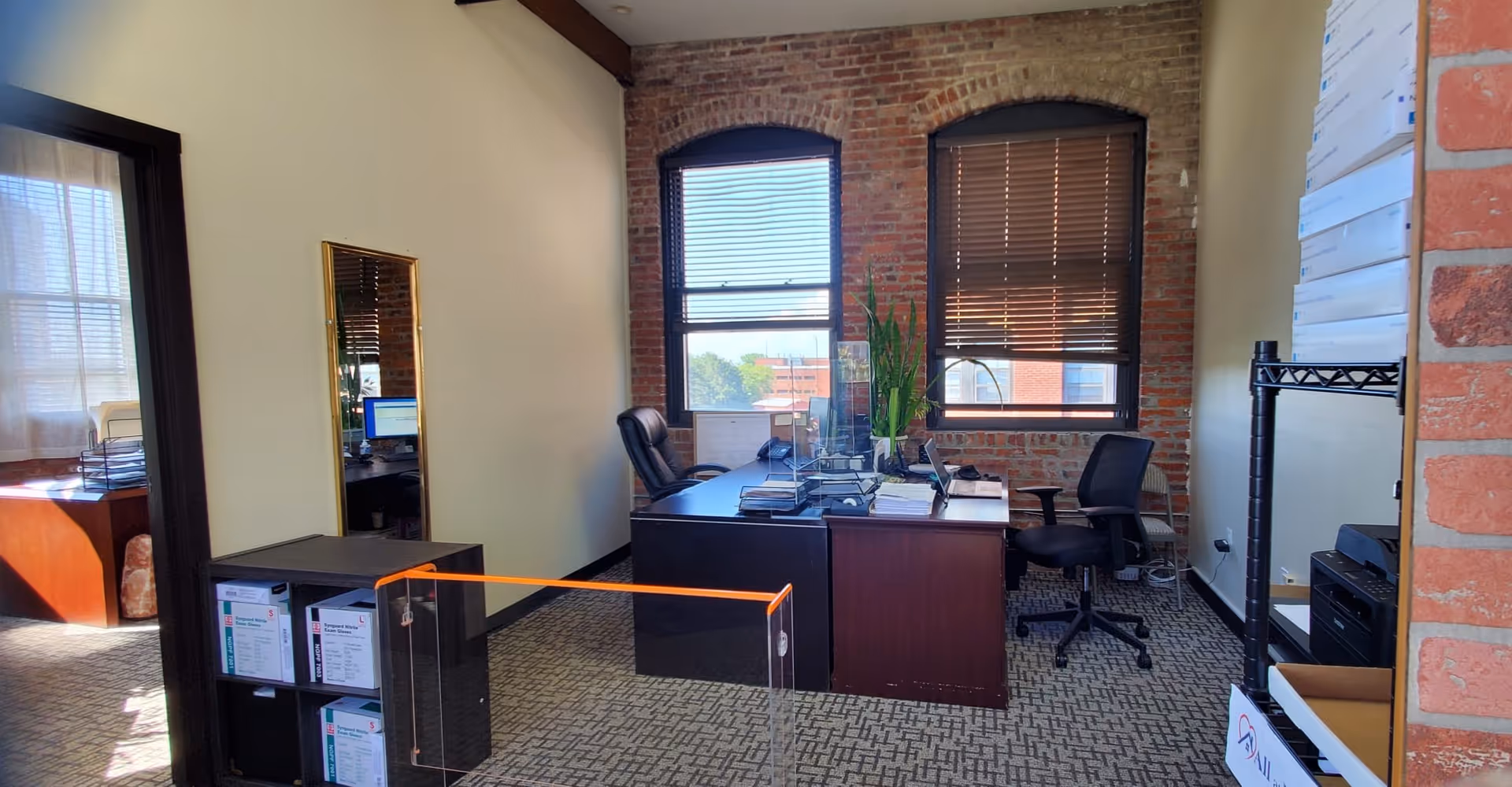 Office workspace with brick wall, two windows, desk, computer, plants, and a glass barrier