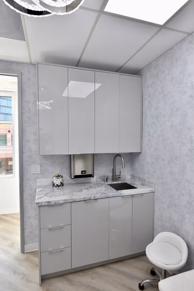 Modern gray kitchenette with marble countertop, sink, upper cabinets, and a white wheeled chair.