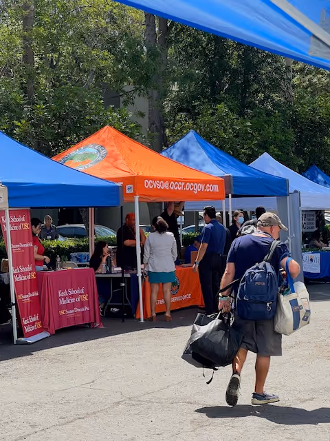 Outdoor community fair with bright tents, tables, and people visiting informational booths.
