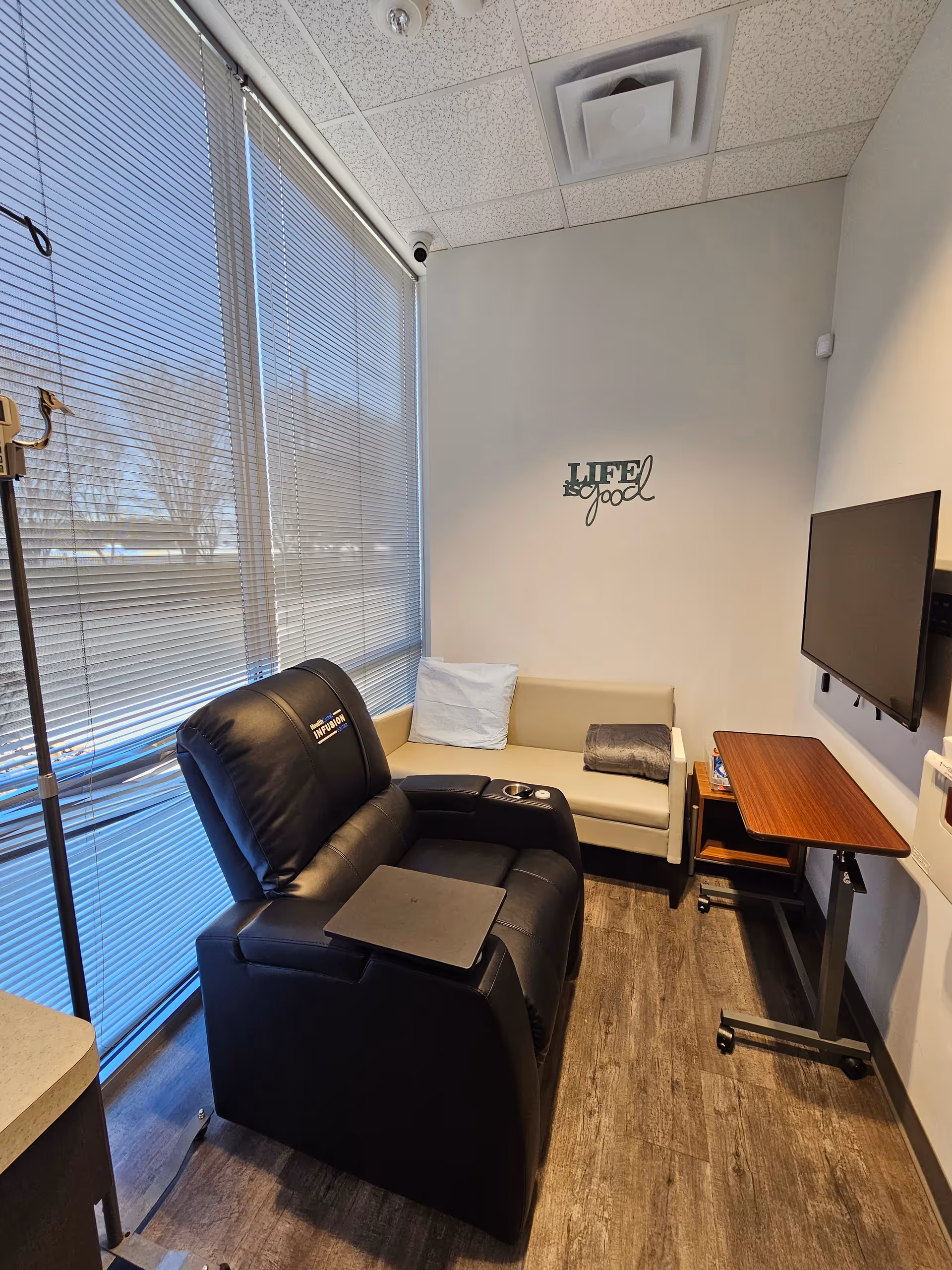 Infusion lounge with a black recliner, beige sofa, TV, and large window blinds.