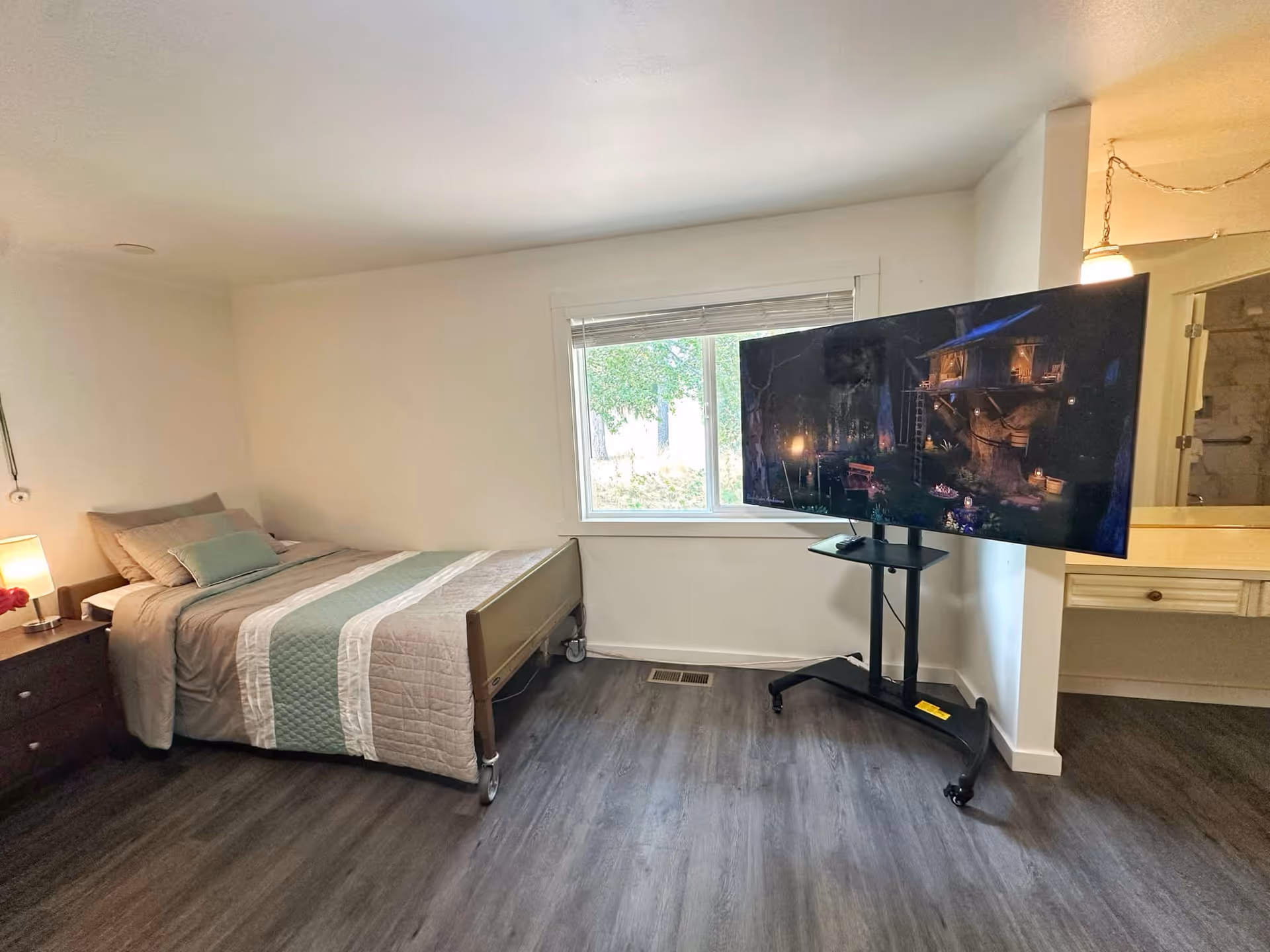 Bedroom with a bed, nightstand, lamp, window, and a large TV on a rolling stand