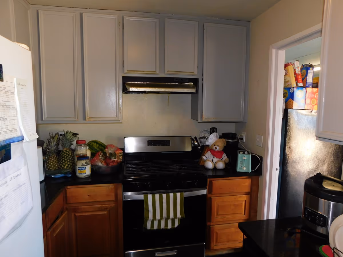 Compact kitchen with white cabinets, stainless stove, fridge, fruit basket, and stuffed bear on counter.