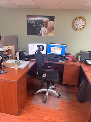 Office workspace with dual computer monitors, chair, desk, clock, and framed photo on wall.