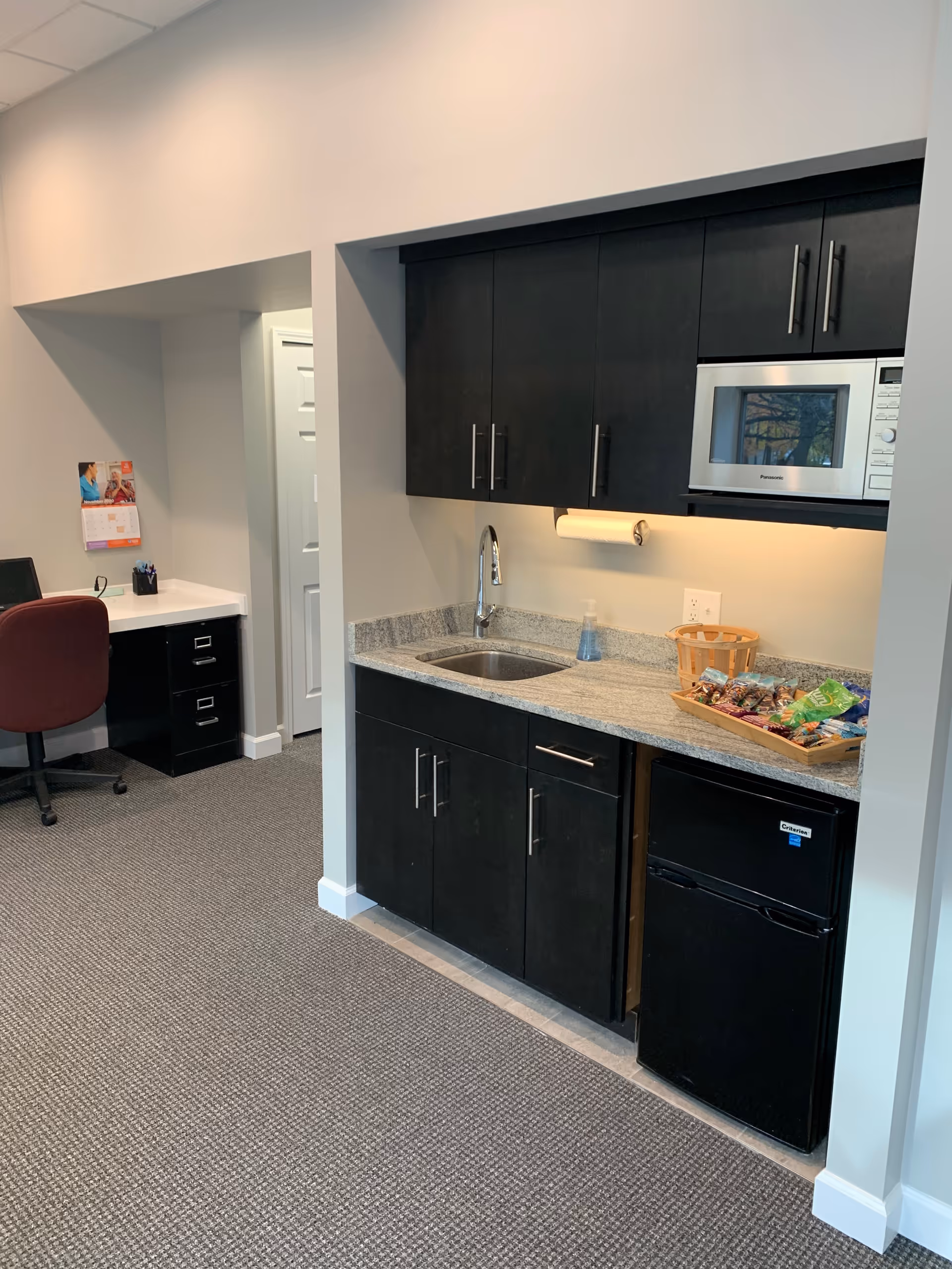 Compact kitchenette with dark cabinets, sink, microwave, mini-fridge, and desk area nearby.