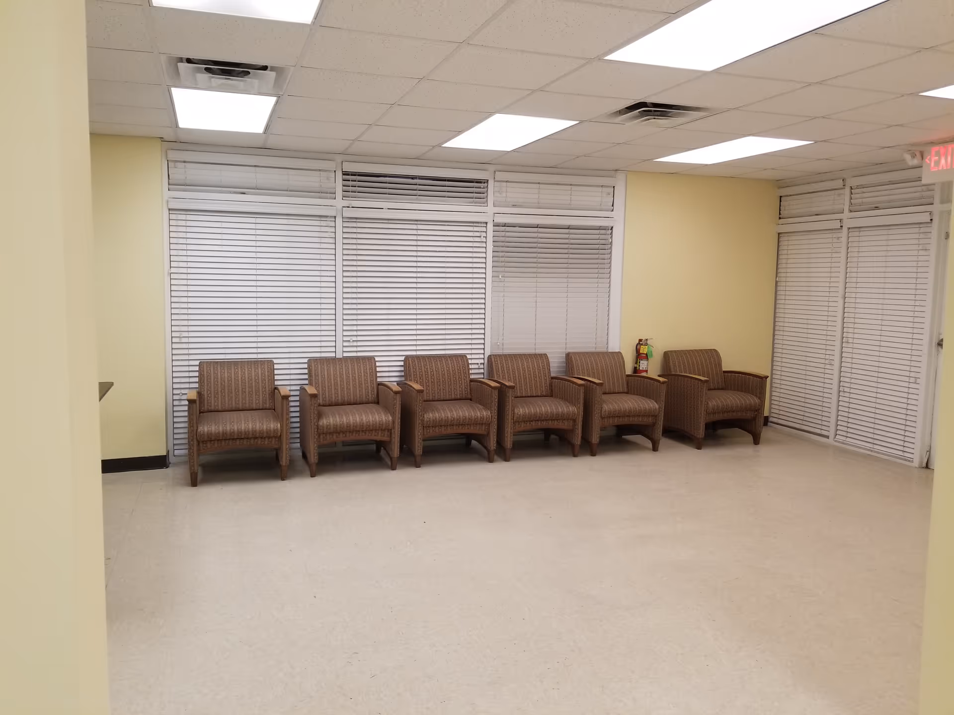 Row of brown chairs along a beige hallway with large blinds and a fire extinguisher.