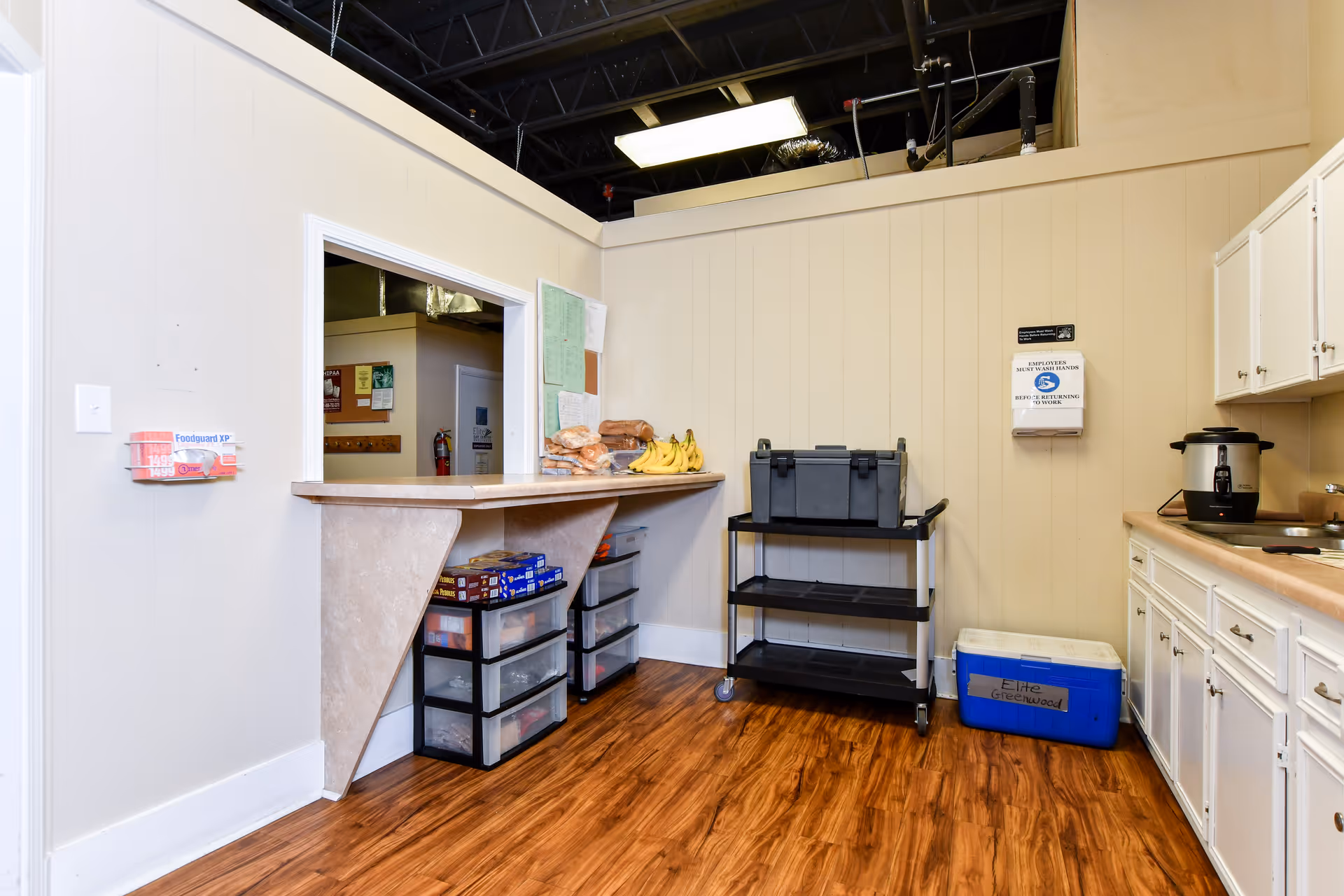 Interior kitchenette in a senior living facility with counter, cabinets, cart, and appliances.