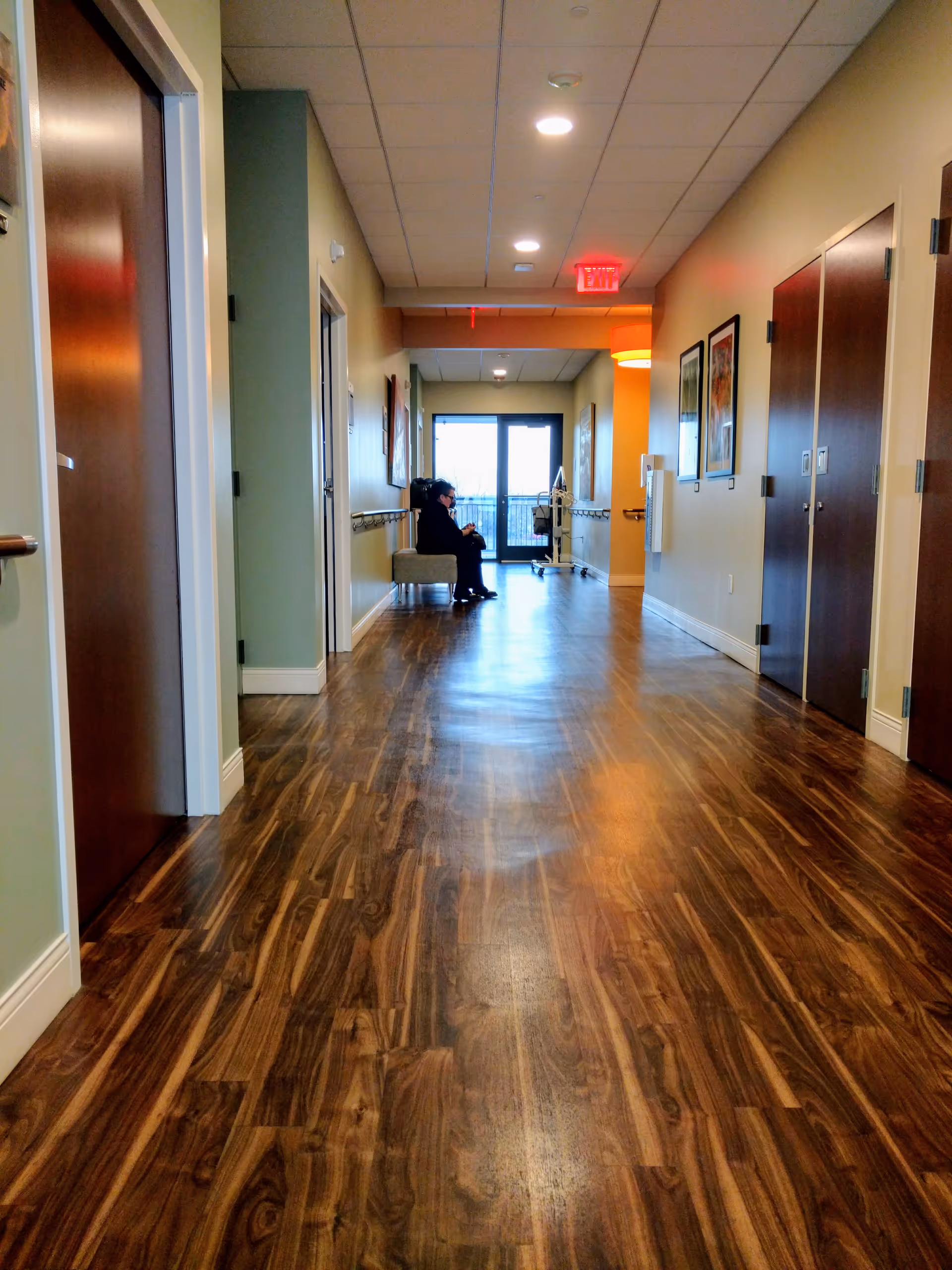 A long interior corridor of a senior living facility with a seated resident near the end.