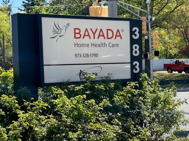 Outdoor BAYADA Home Health Care sign beside street with green bushes and vertical 3-8-3 numbers.