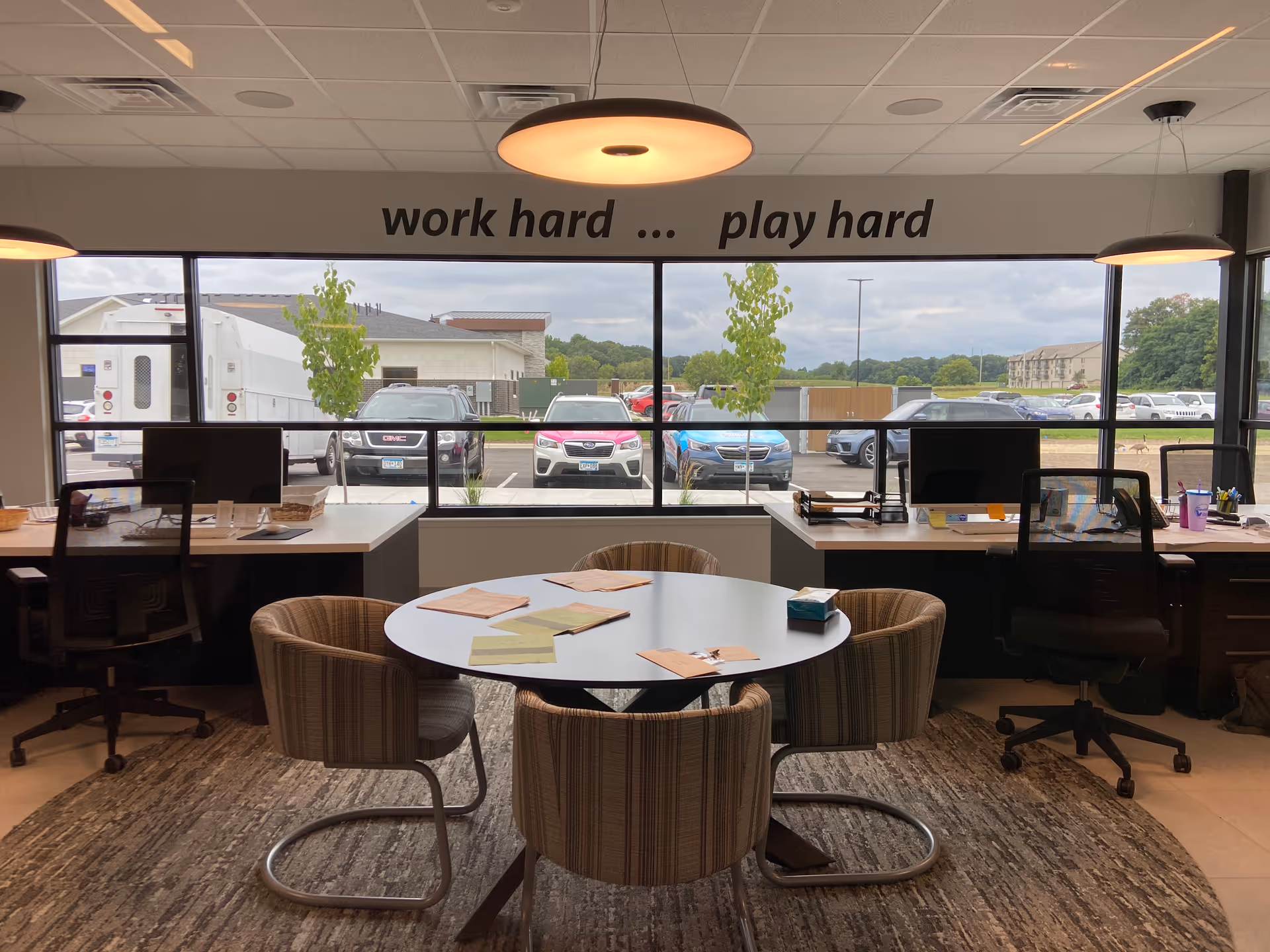 Office-style common area with round table, desks, computers, and large windows.