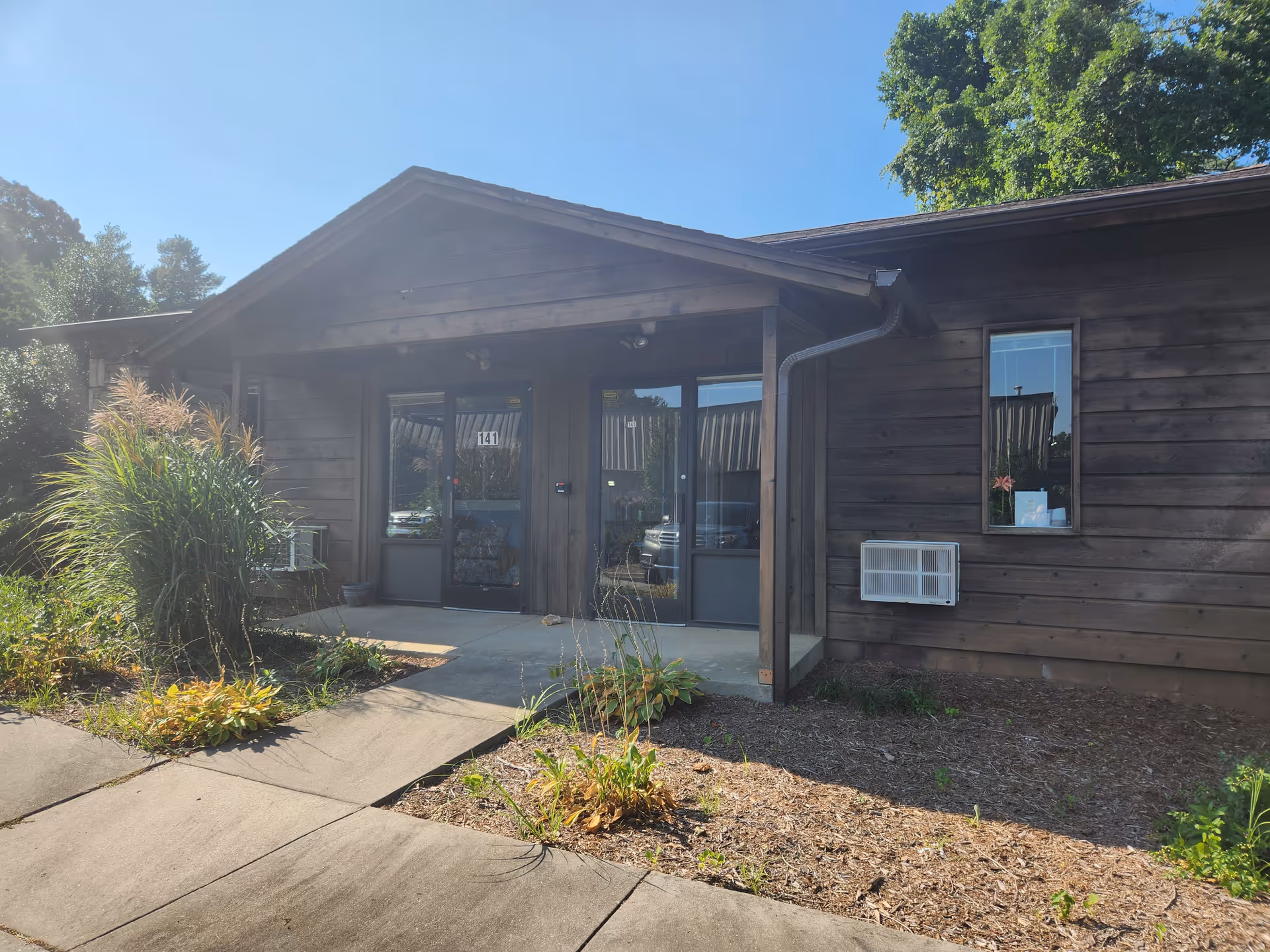 Wooden facility entrance with glass doors, walkway, and landscaping on a sunny day.