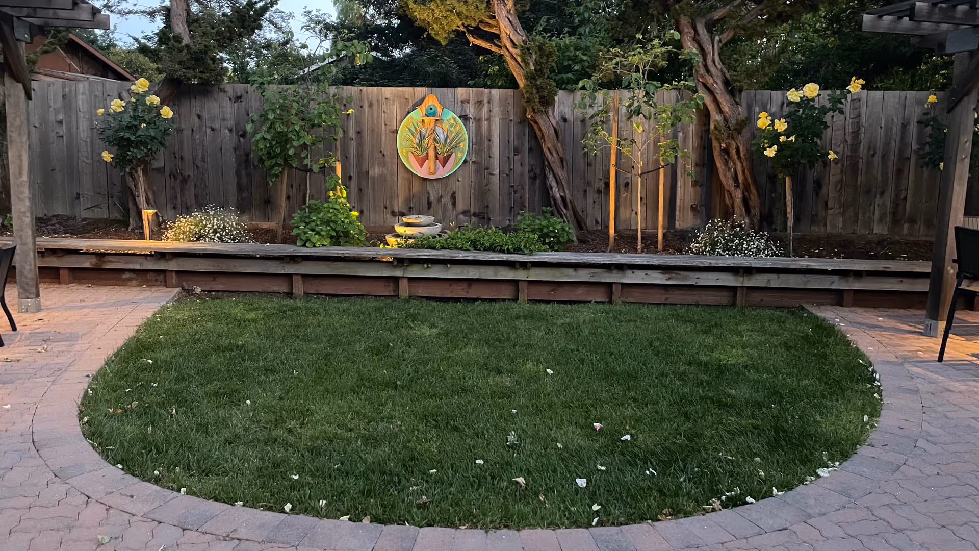 Backyard courtyard featuring a circular lawn, raised planting beds, fence, and flowering plants.