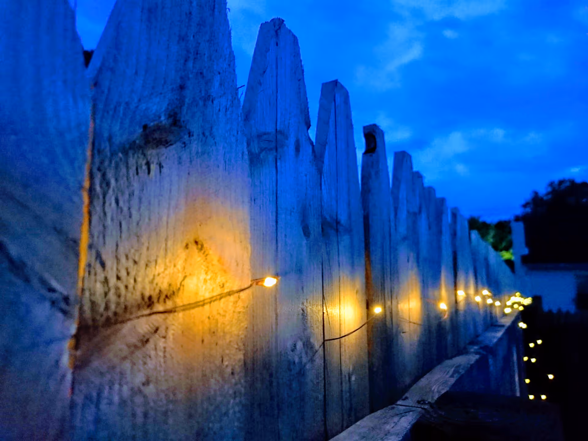 Blue dusk exterior view of a wooden fence lined with warm string lights.