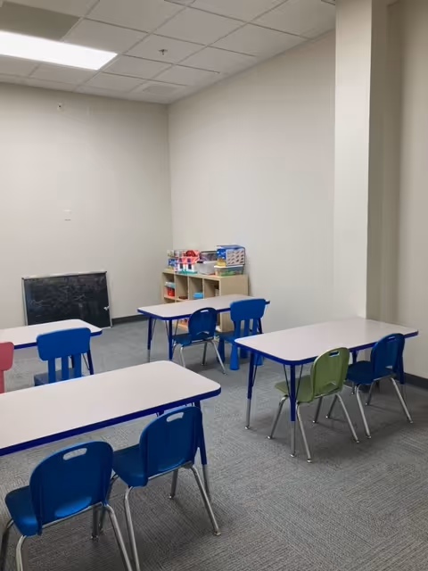 A bright indoor activity room with small tables, blue chairs, and a toy shelf.