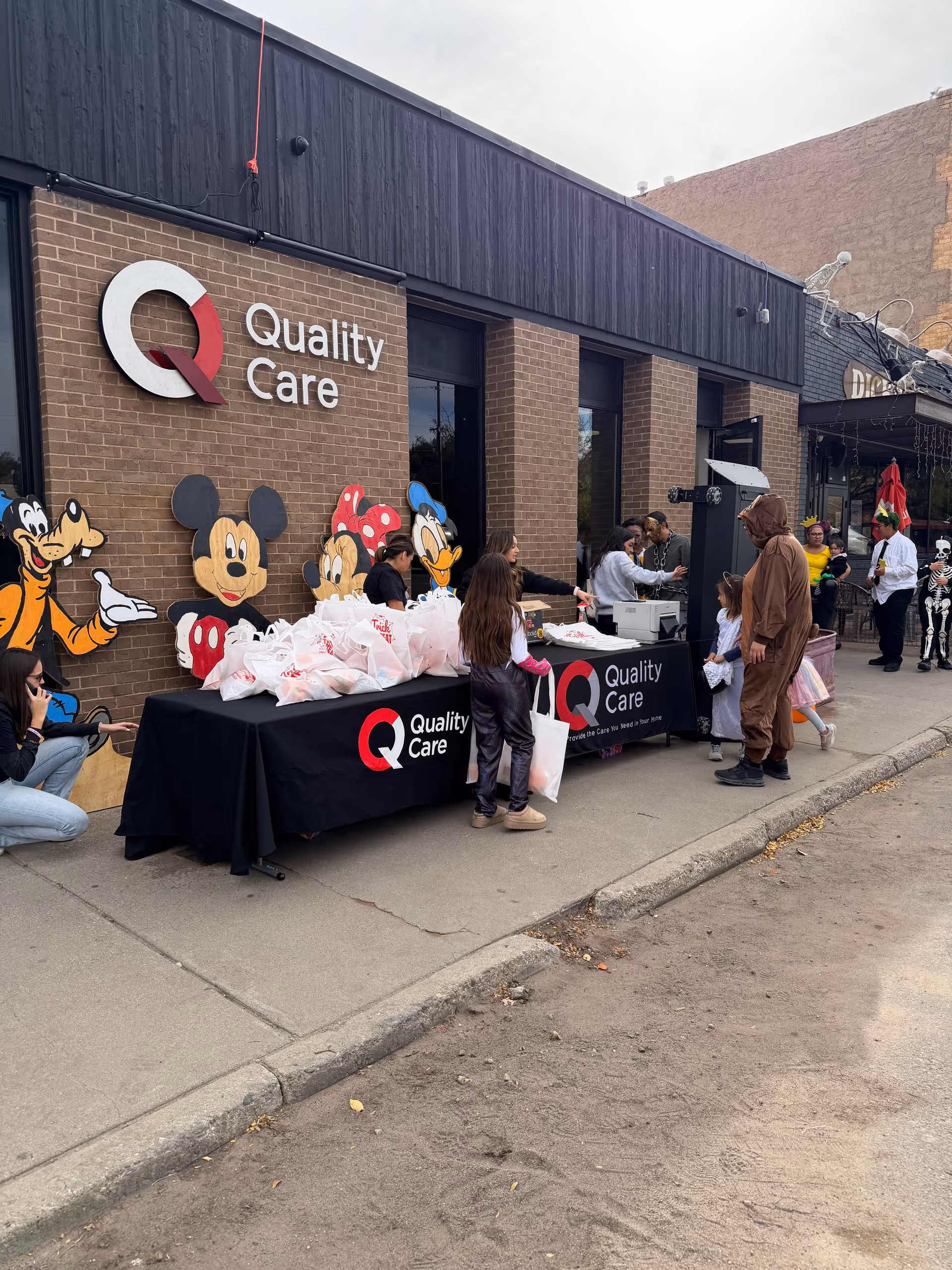 Outdoor event in front of Quality Care with tables, bags, and people.