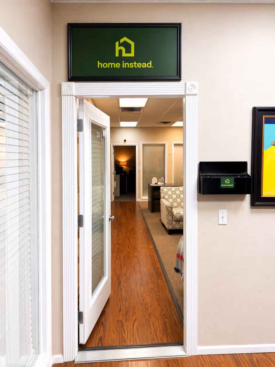 Interior hallway of a Home Instead facility with sign above doorway and seating area beyond.