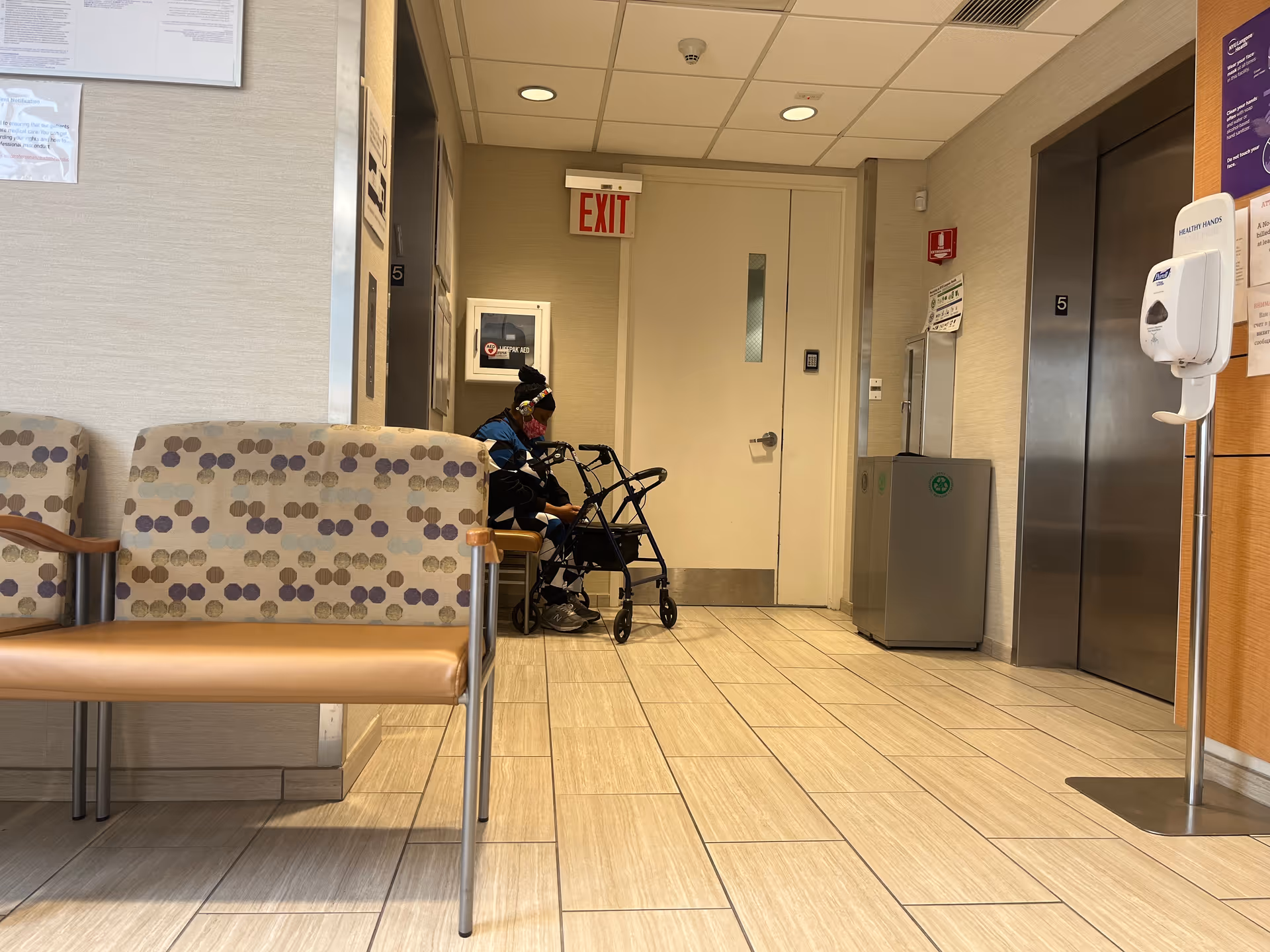 Interior hospital-like hallway with patterned seating, a person with a walker near an elevator.