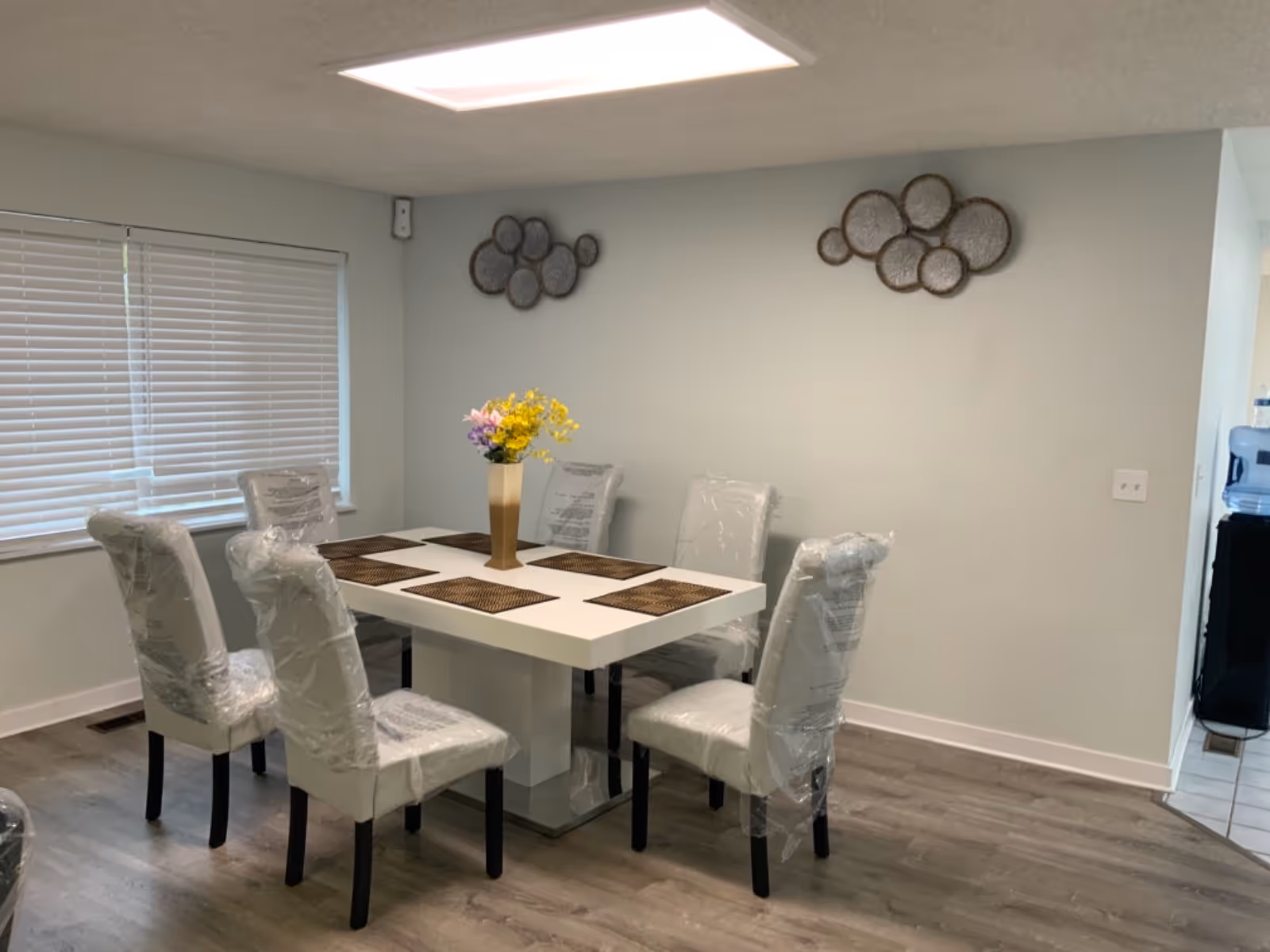 Dining room with a white table, six plastic-wrapped chairs, and a flower centerpiece.