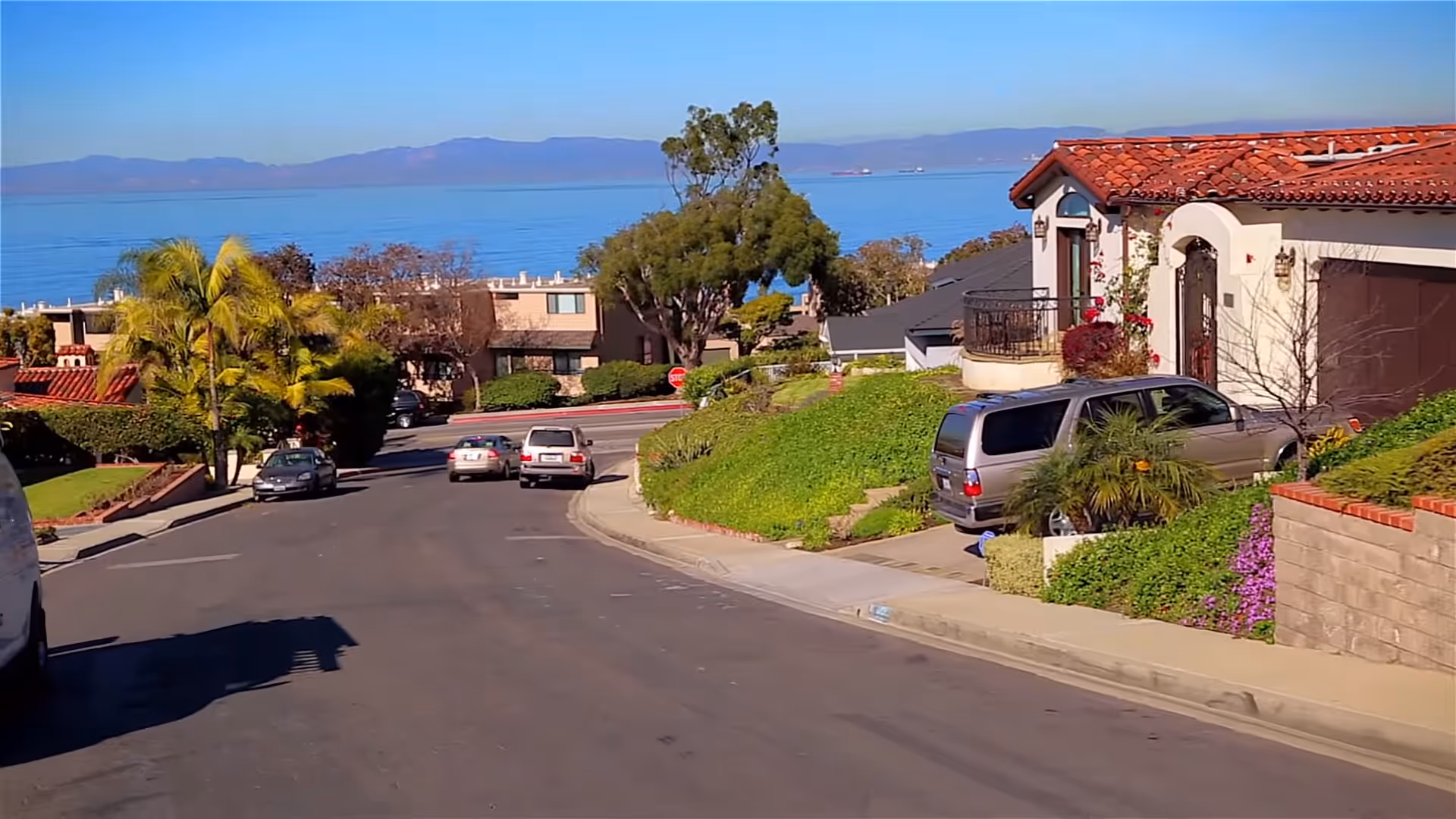 Sunny coastal street with houses, cars, trees, and distant ocean view.