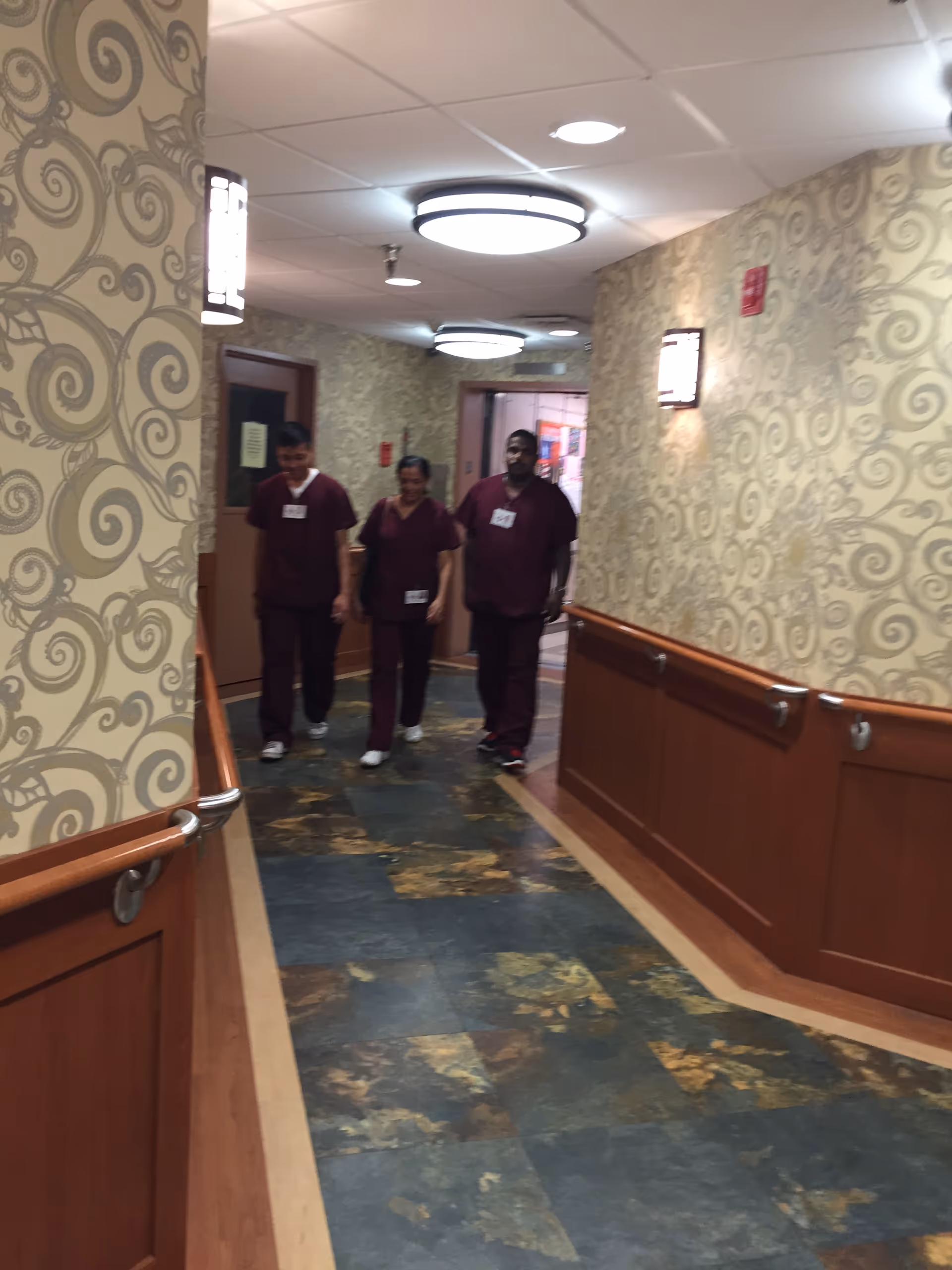 Three staff members in maroon scrubs walk down a patterned hallway inside a senior living facility.