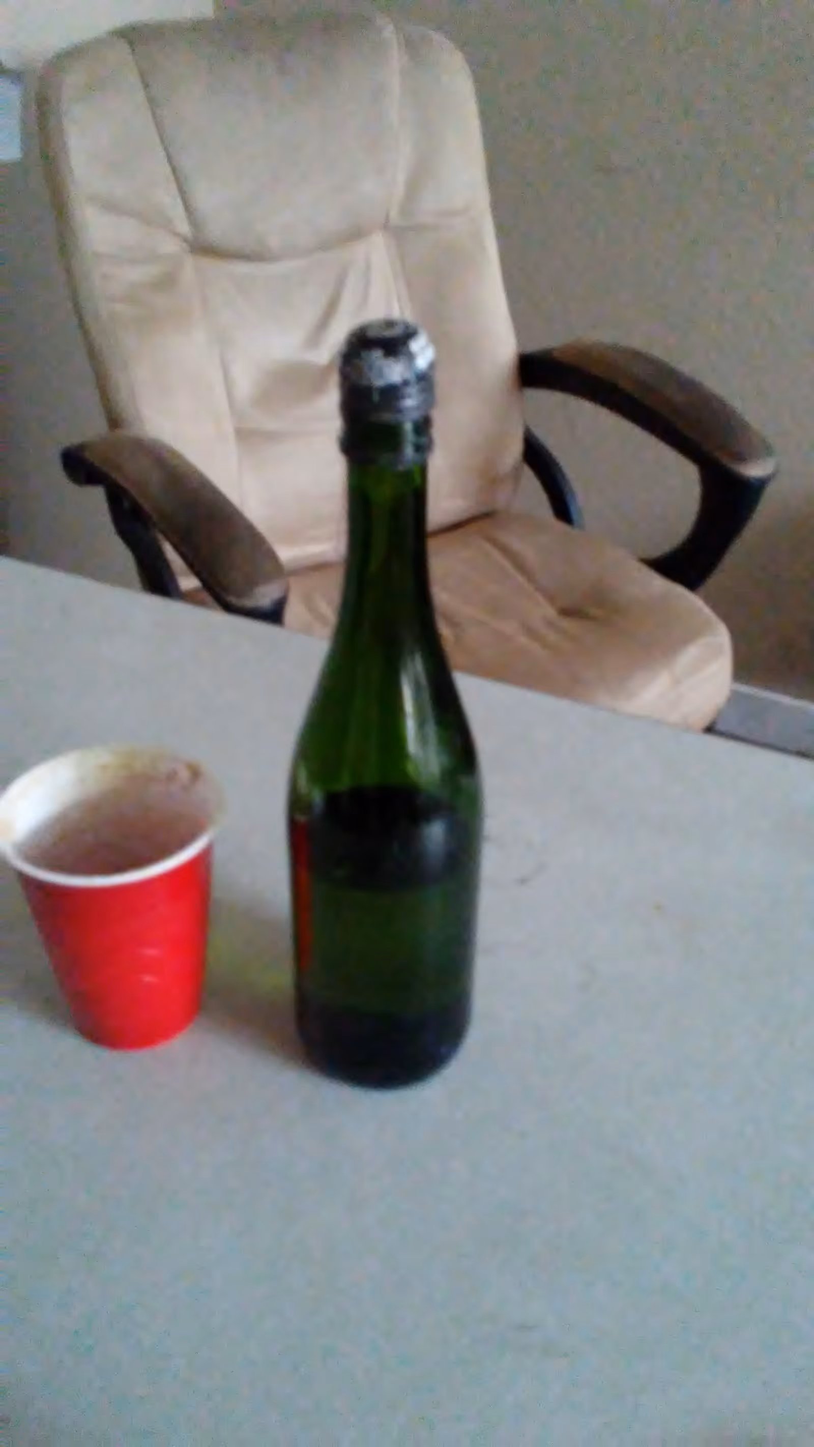 Bottle and red cup on a table with a beige chair in a room.
