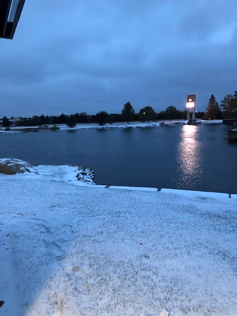 Snow-covered lakeside scene at dusk with distant lit sign and trees.