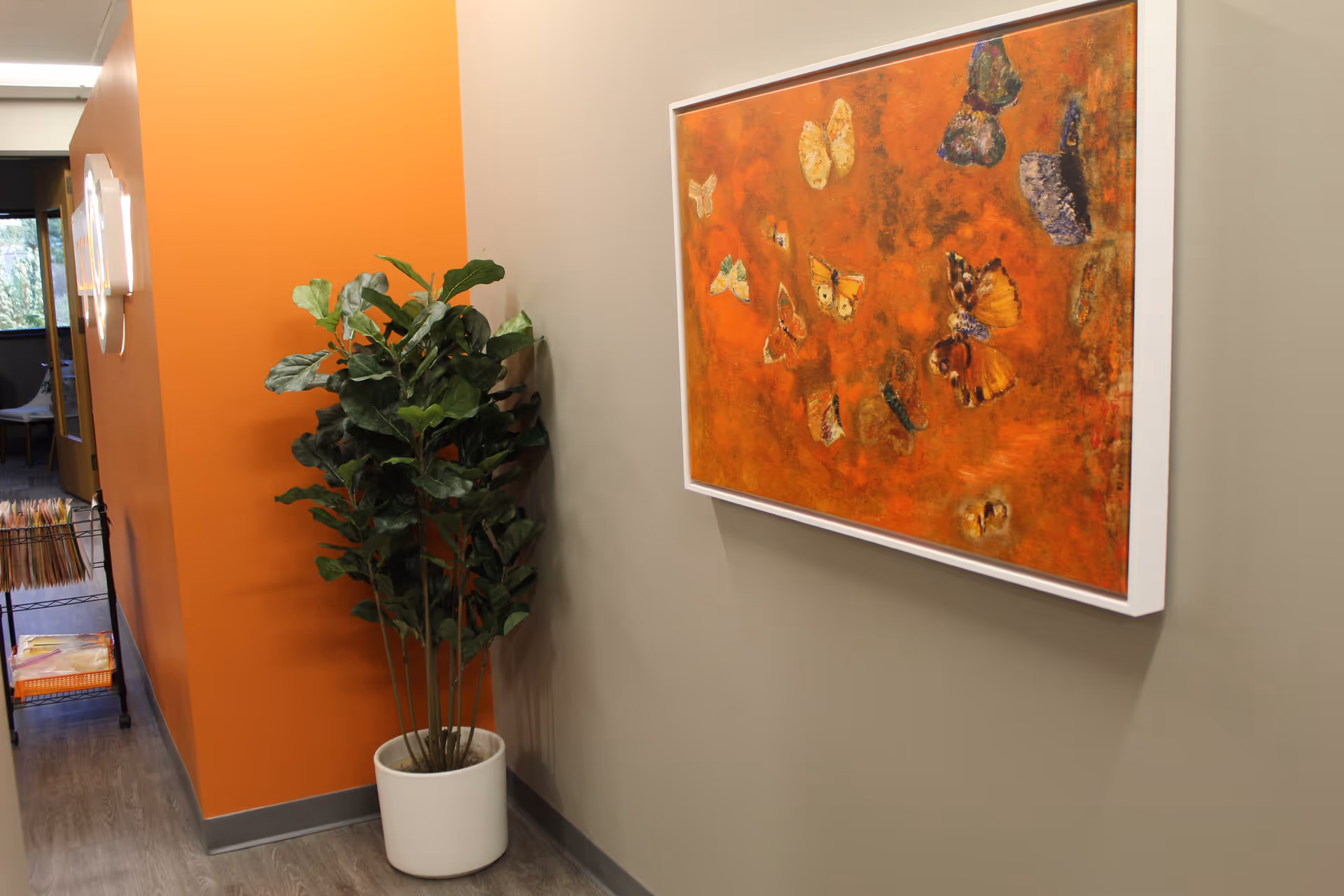 Indoor hallway with orange wall, a tall potted plant, and a large framed butterfly painting.