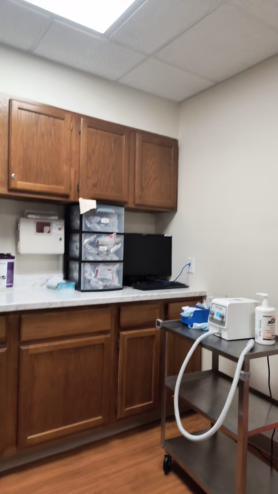 Medical supply area with wooden cabinets, countertop, computer monitor, and rolling cart.