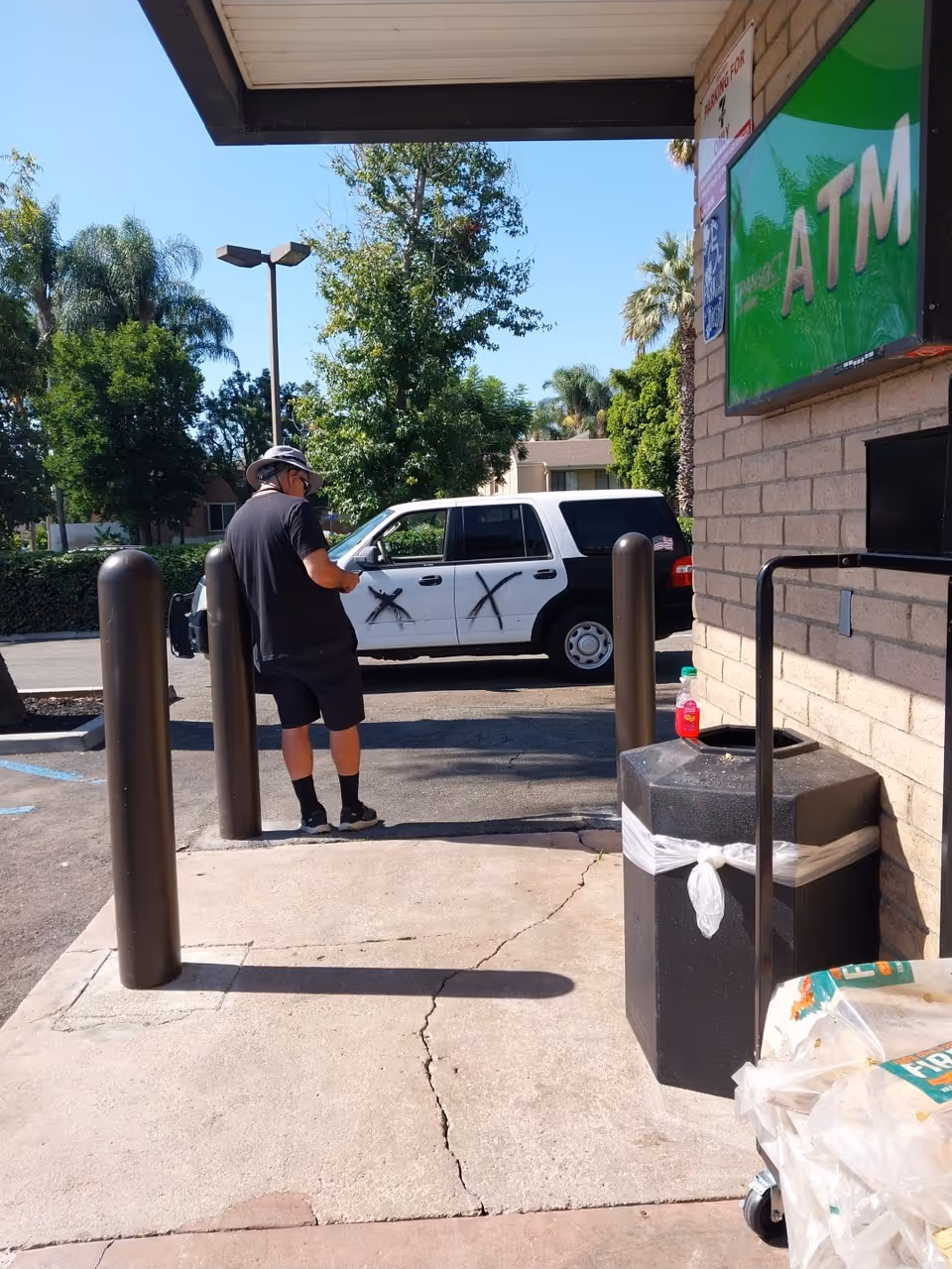 Man stands outside a brick building near an ATM, with a white SUV in the background.