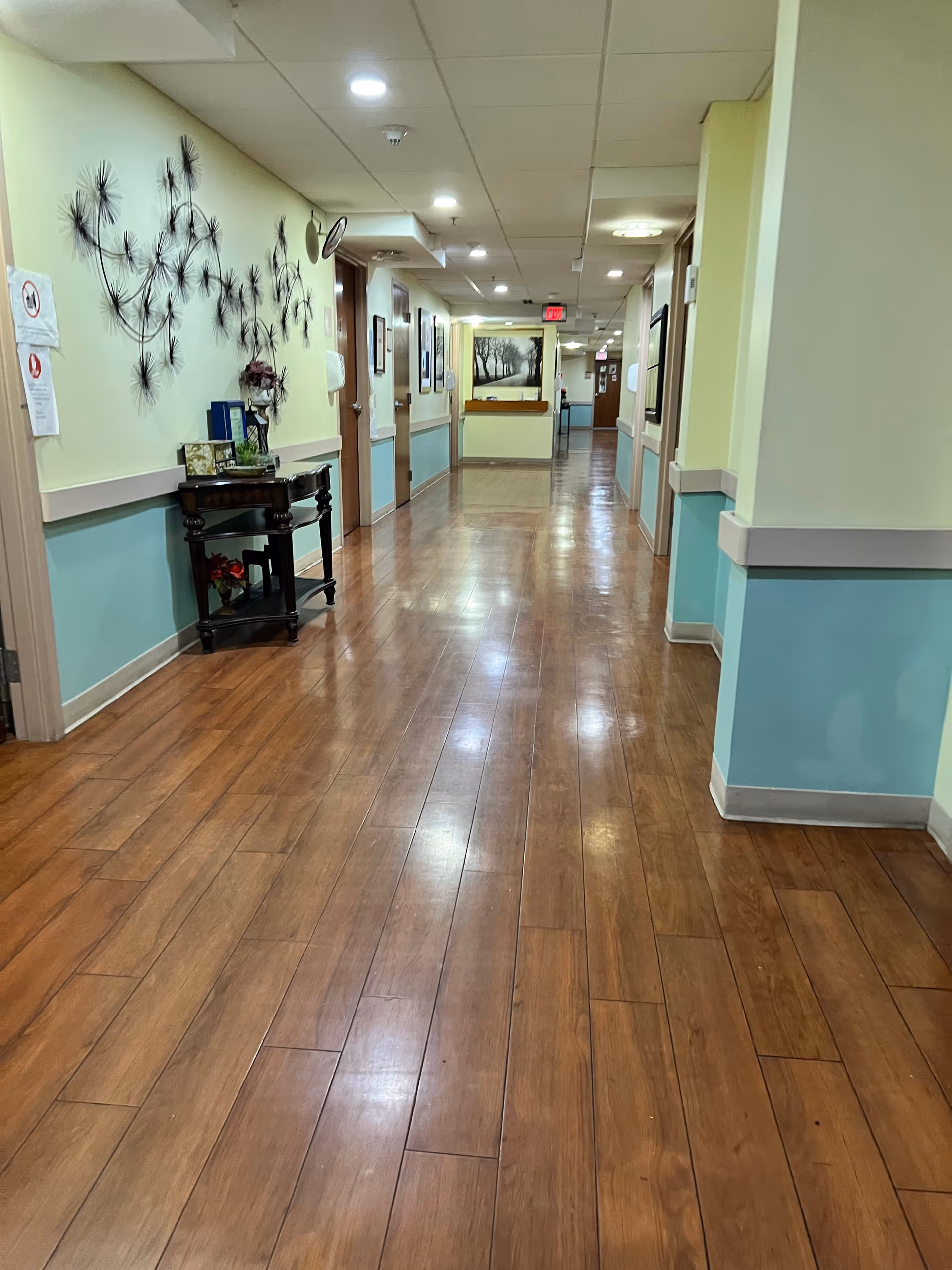 Long, well-lit hallway in a senior living facility with wood flooring and wall art.