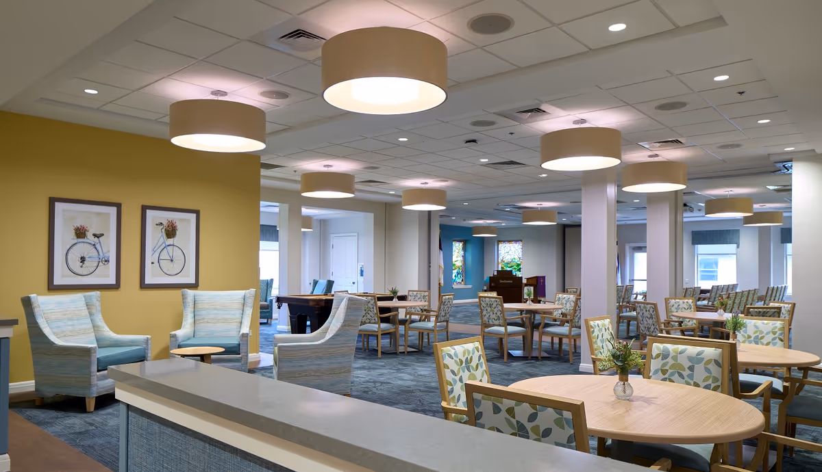 Spacious senior living common area with round tables, cushioned chairs, and ceiling lights.