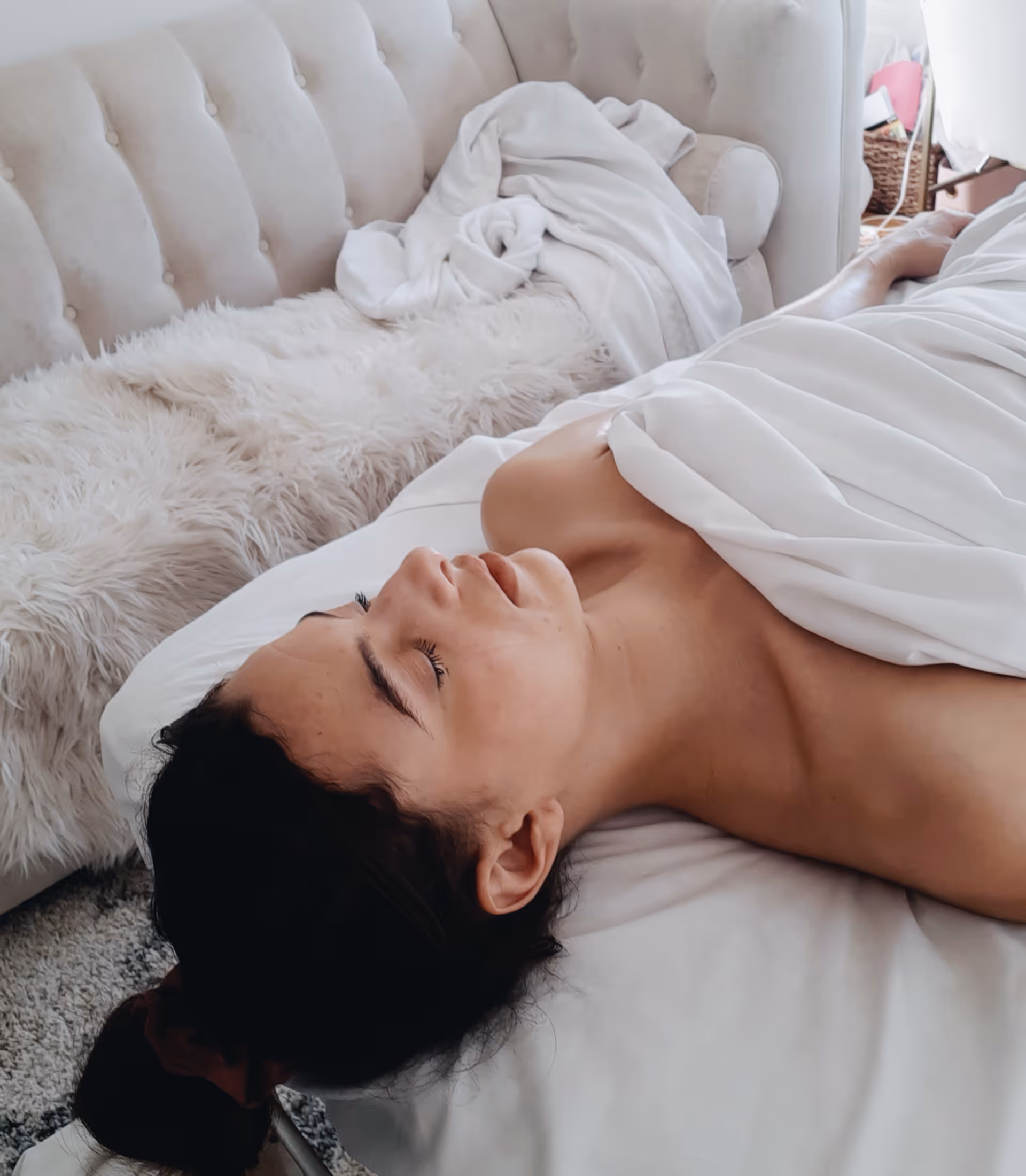 A person lying in white sheets on a tufted bed in a cozy bedroom.