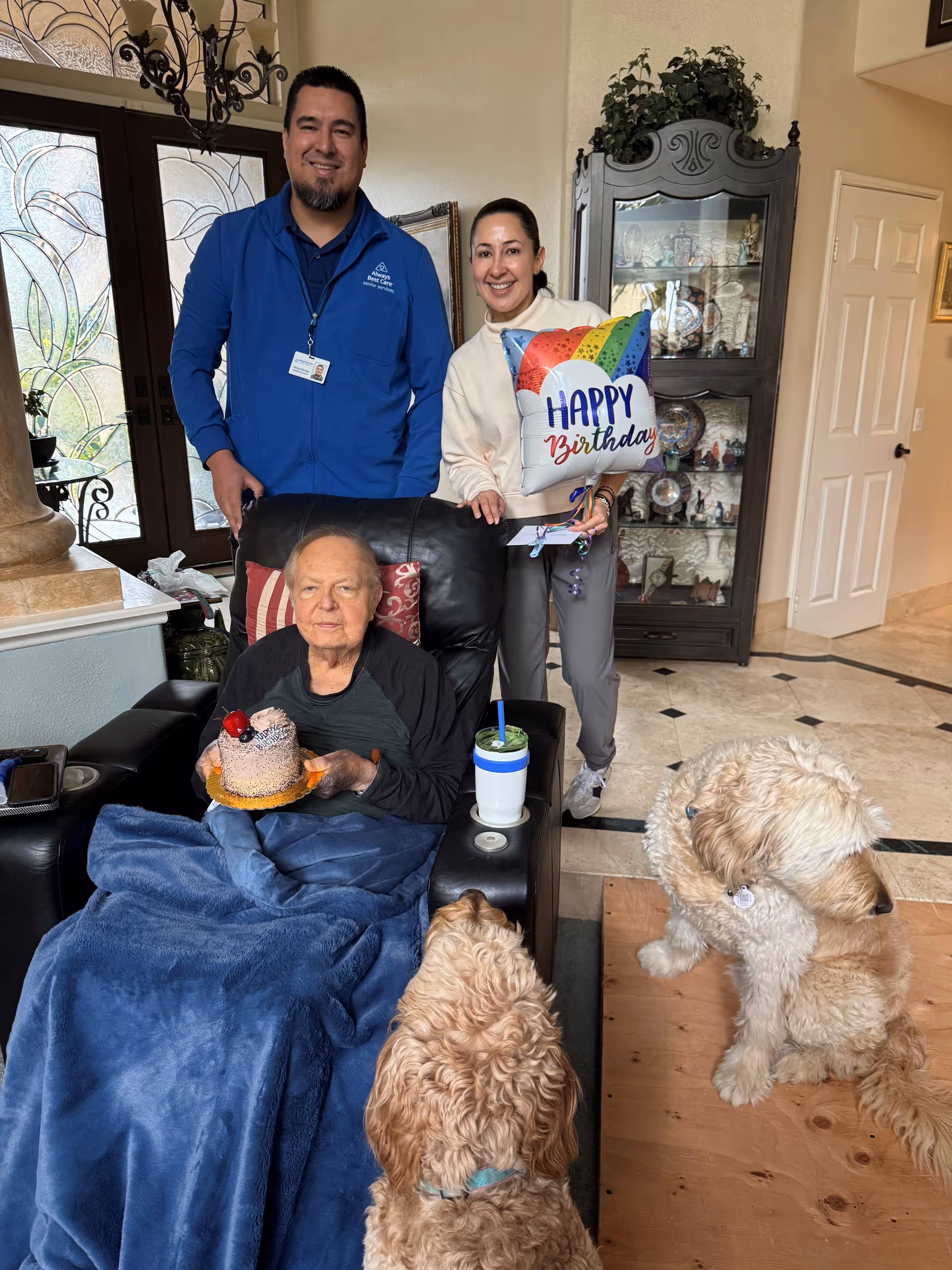 Senior resident celebrates birthday with caregivers in a living room, holding cake; dogs nearby.