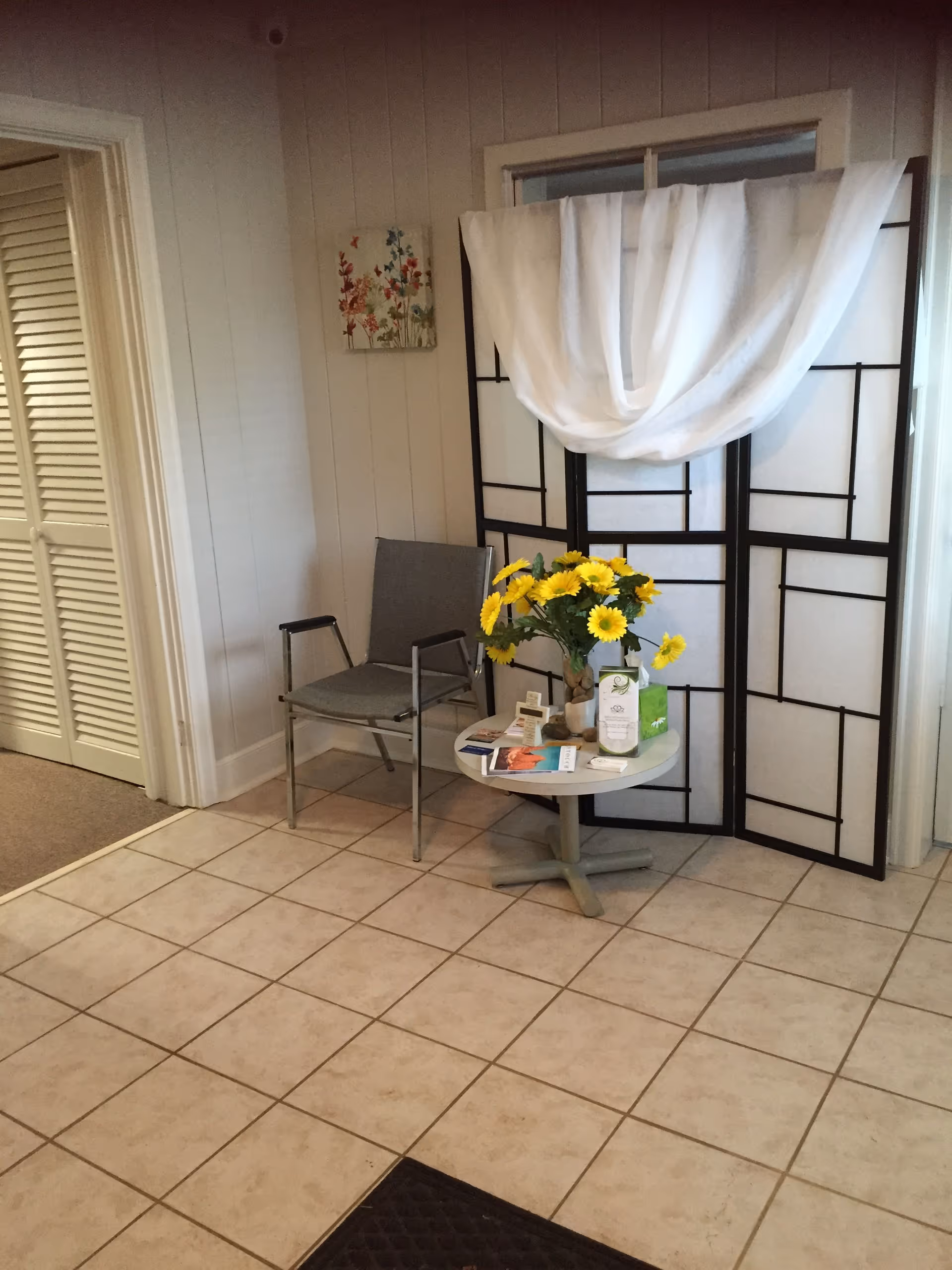 Small lobby corner with a chair, round table, sunflowers, and a folding screen.