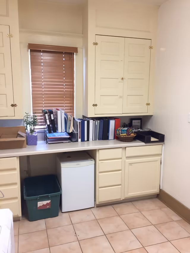 Cream cabinets and counter with binders, a small fridge, and a window with blinds.