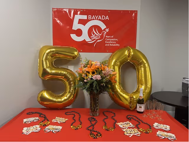 Indoor celebration display with gold '50' balloons, BAYADA banner, flowers, and table setup.