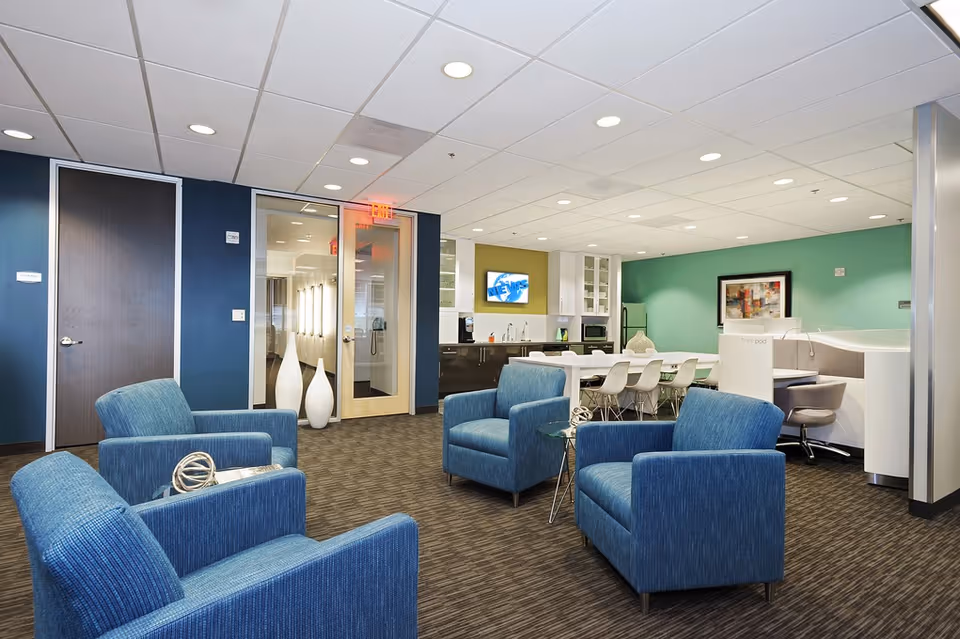 Senior living facility lounge with blue chairs, a dining table, and a kitchenette.