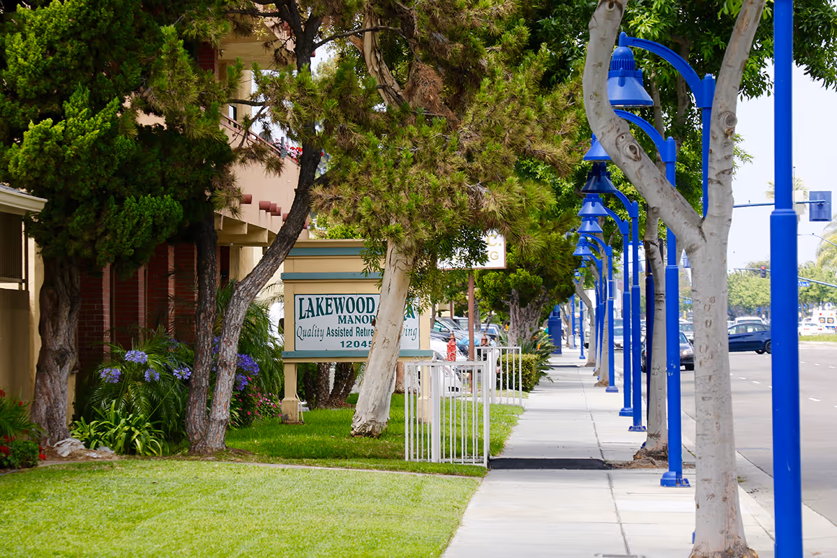 Tree-lined sidewalk outside a senior living facility with blue lamp posts and Lakewood Manor sign