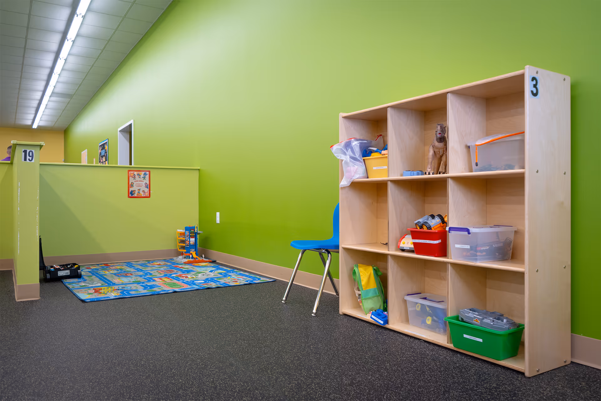 Bright green interior play area with cubbies, bins, a chair, and a colorful rug.