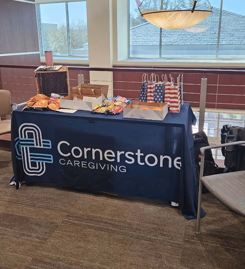 A table with snacks, gift bags, and a banner reading Cornerstone Caregiving inside a facility.