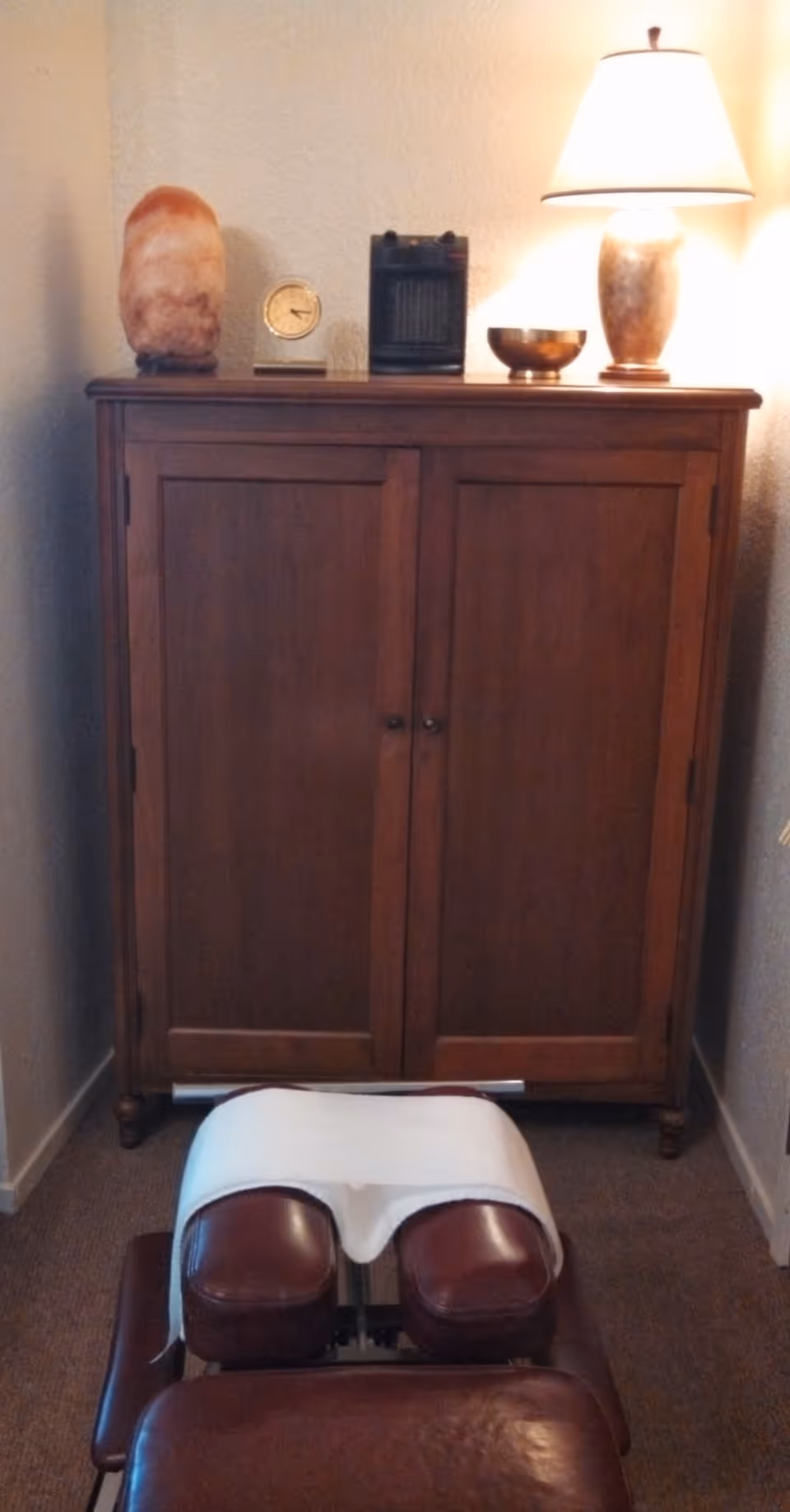 Wooden cabinet with lamp and massage chair in a narrow, softly lit room.