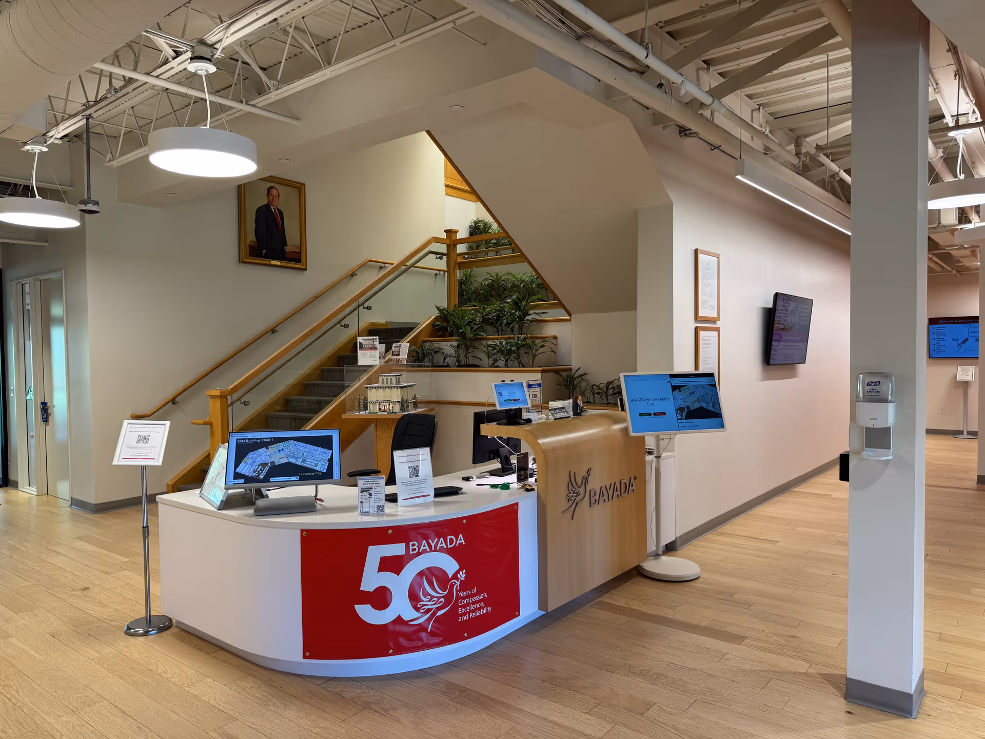 Reception desk with computer monitors in a BAYADA lobby, featuring a staircase and plants.