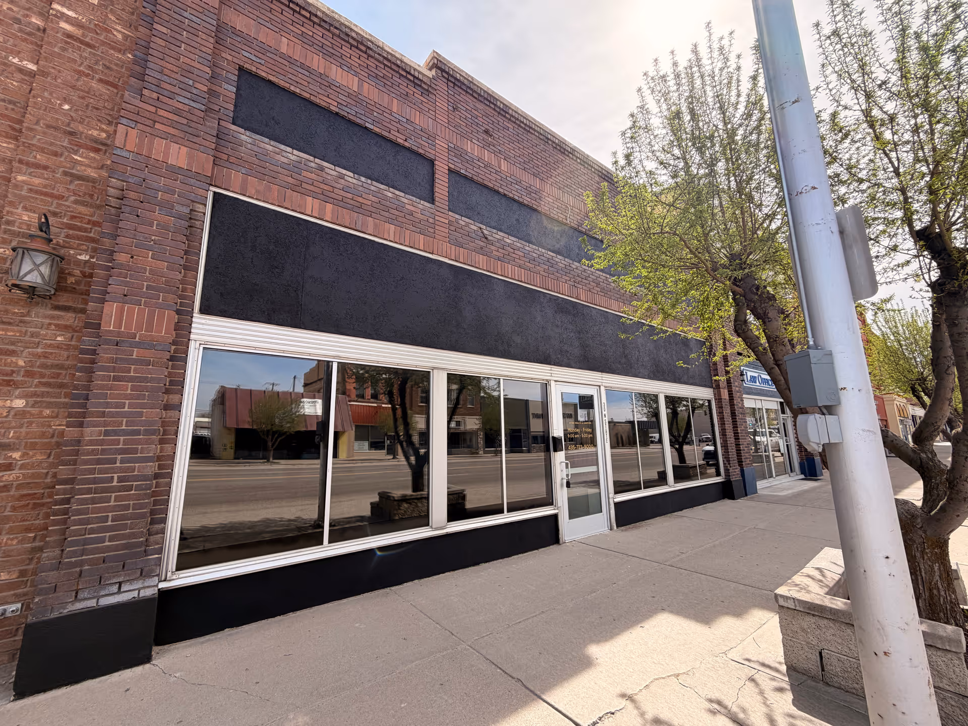 Brick storefront with large windows along a sidewalk on a sunny street.
