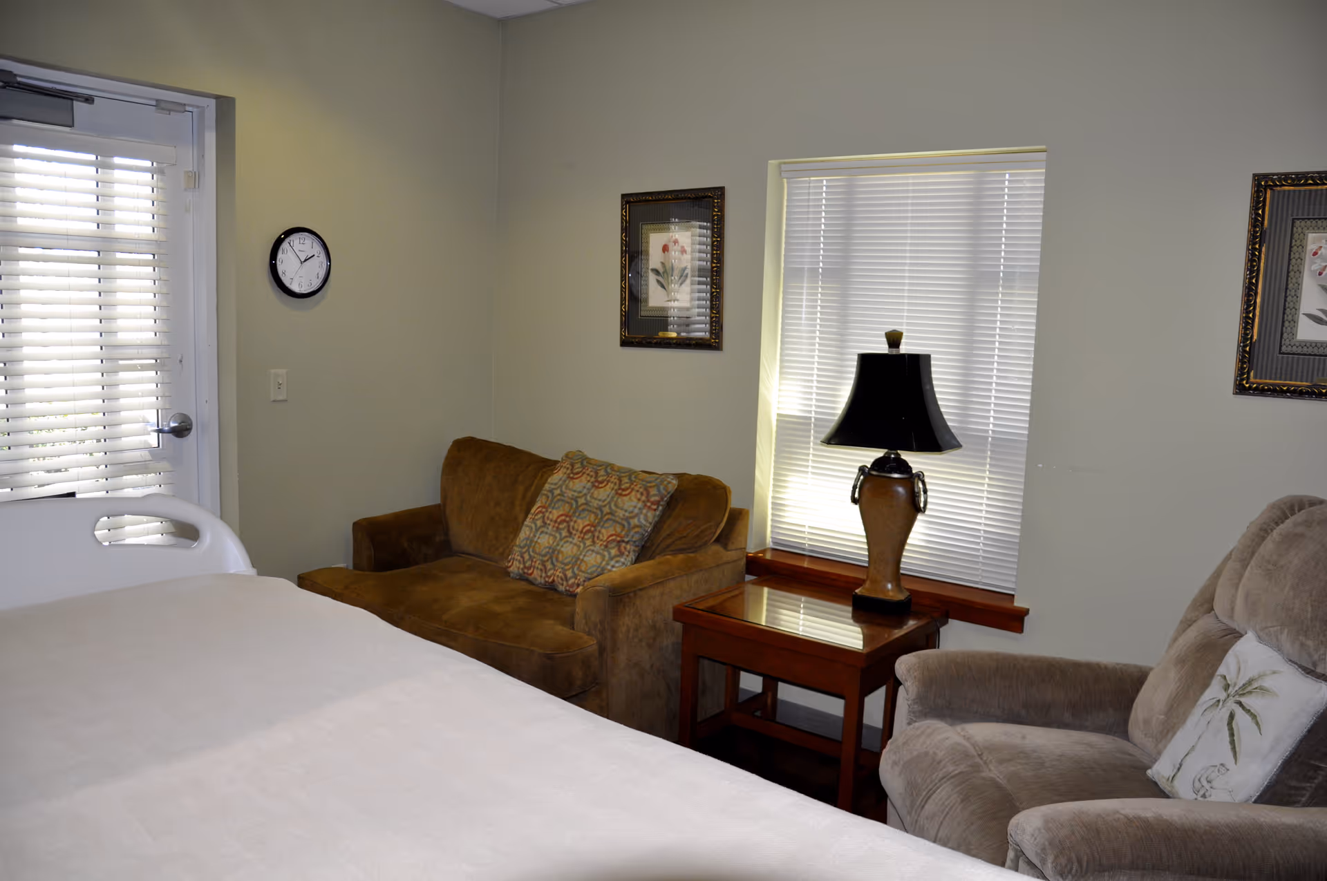 Bedroom in a senior living facility featuring bed, seating, lamp, and window blinds.