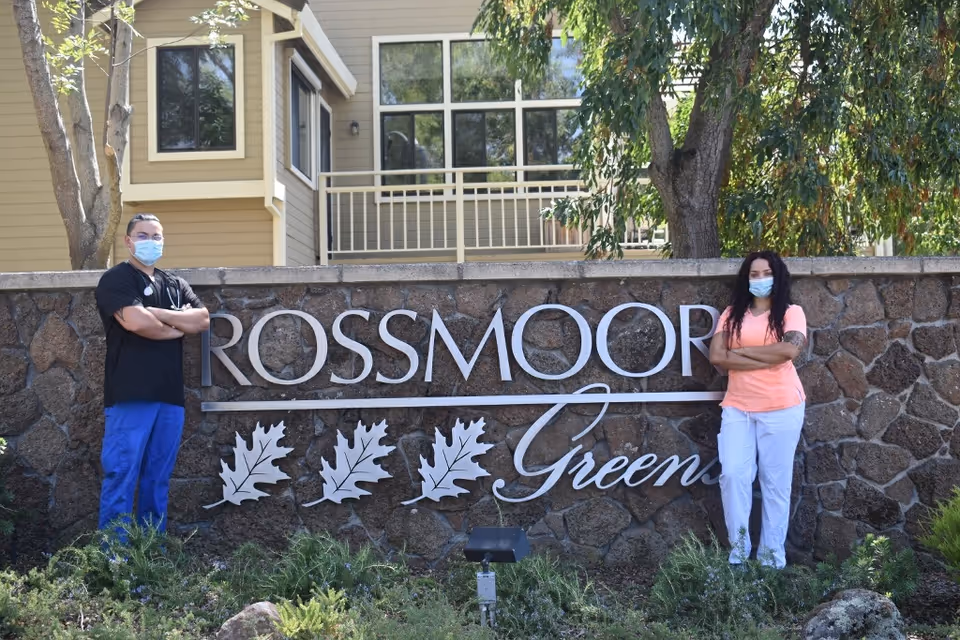 Two masked people stand on either side of a stone sign that reads Rossmoor Green outdoors.