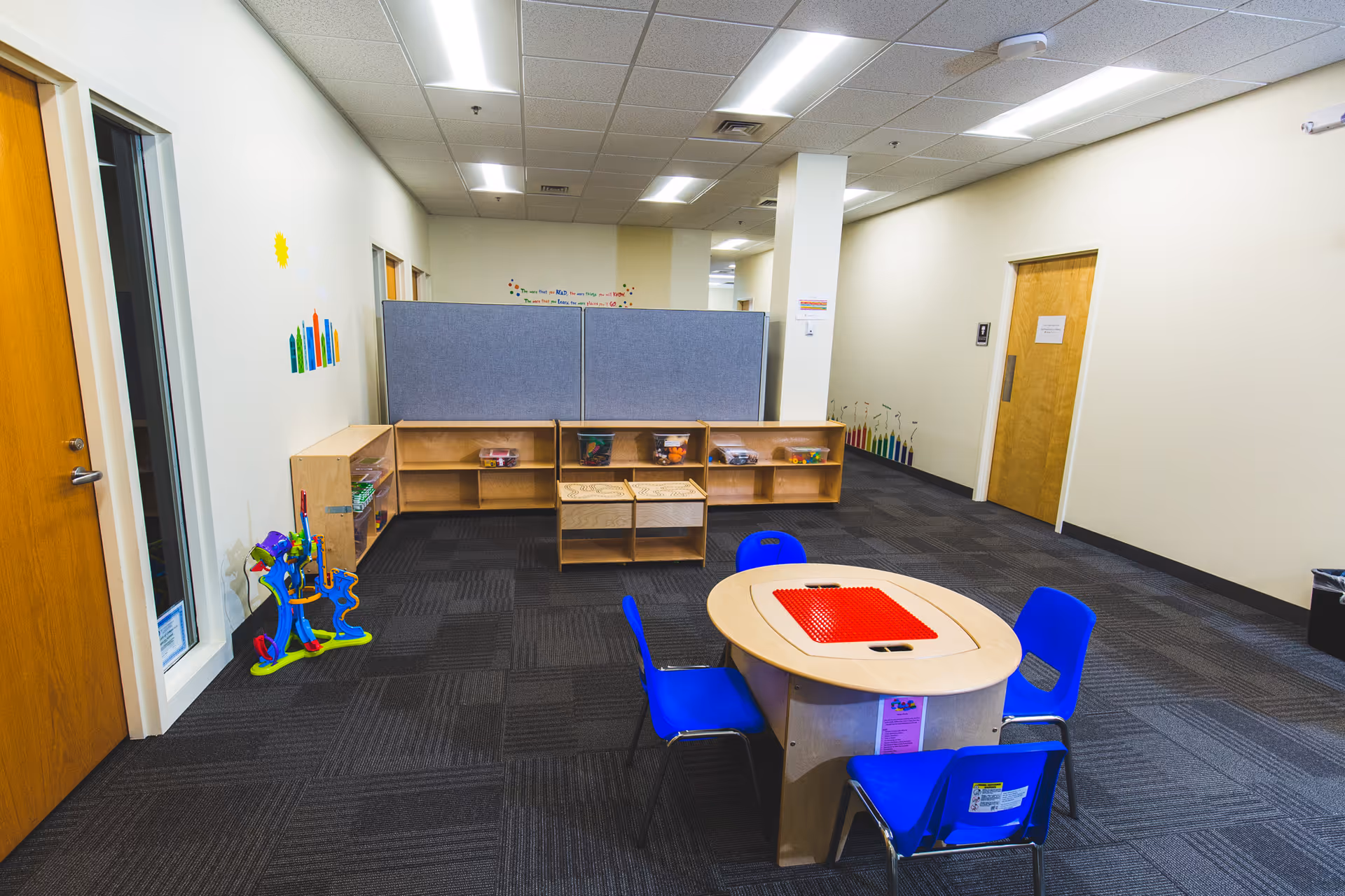 Small round table with a red grid inlay, blue chairs, and toy shelves in a hallway.