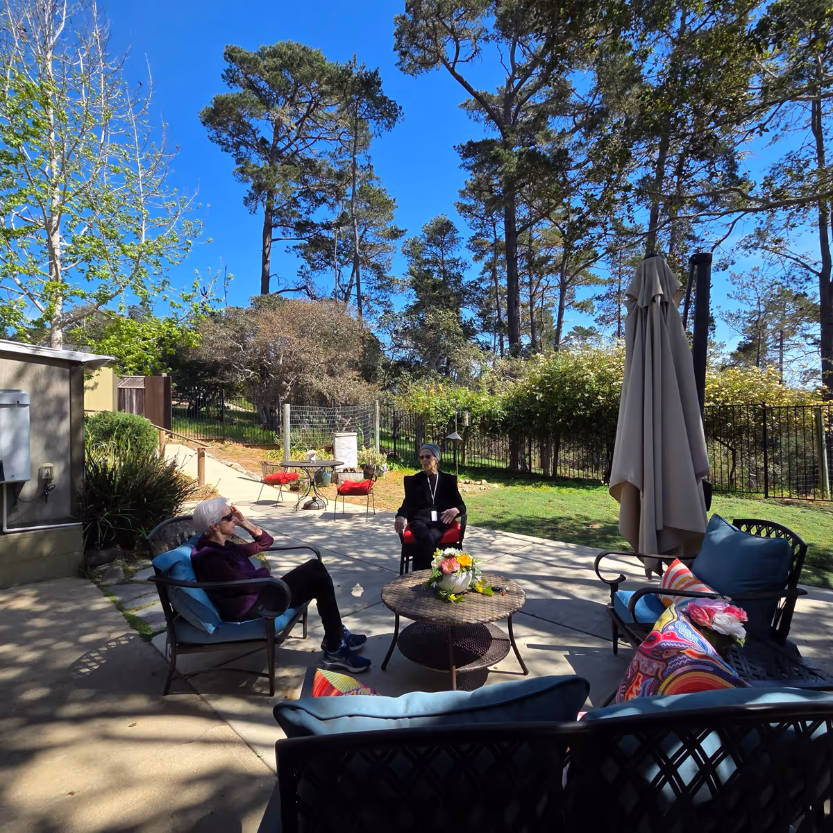 Residents chat on a sunny outdoor patio with seating, tables, an umbrella, and trees.