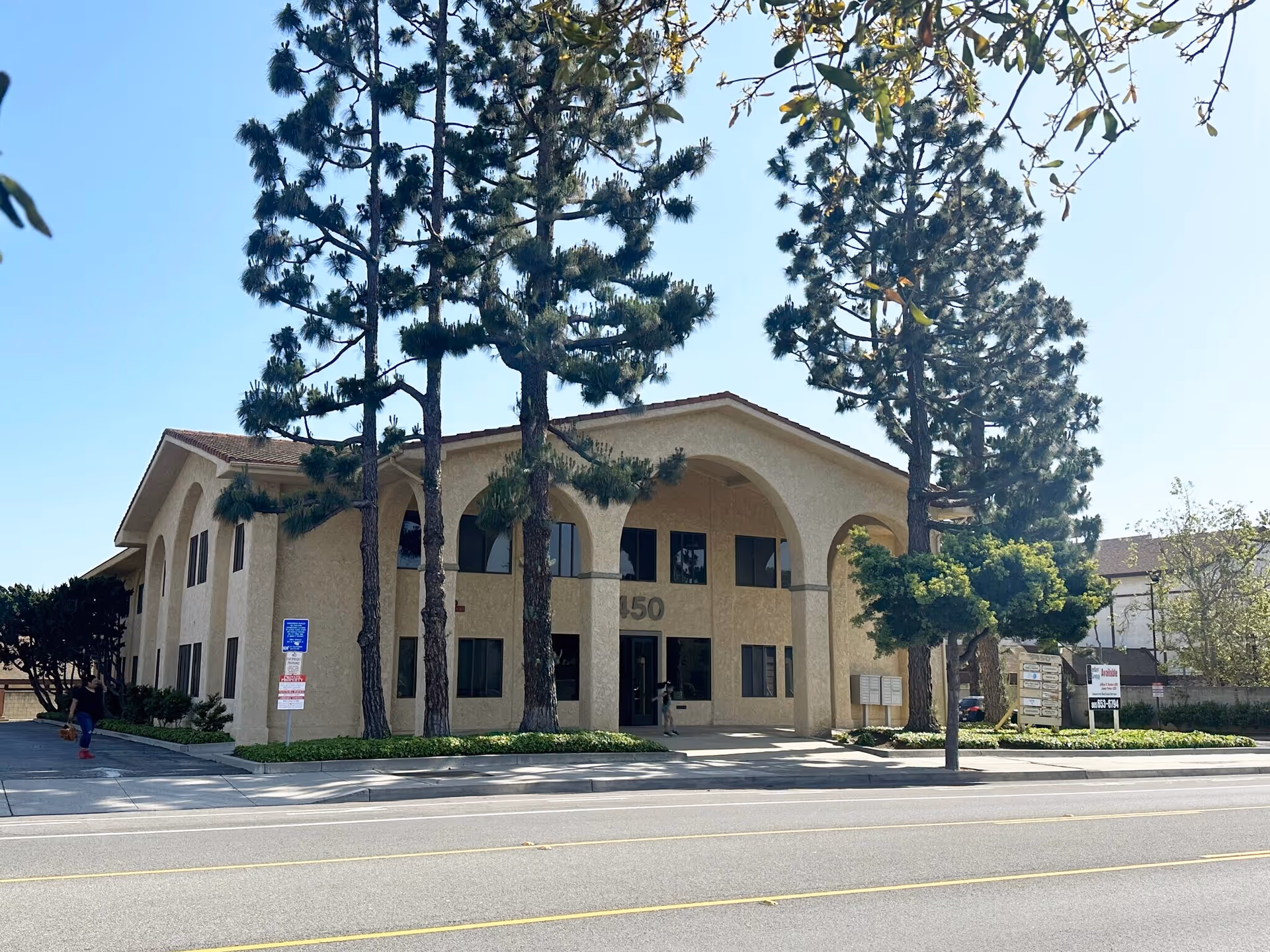 Exterior view of a beige two-story building with arches and tall trees by a street.